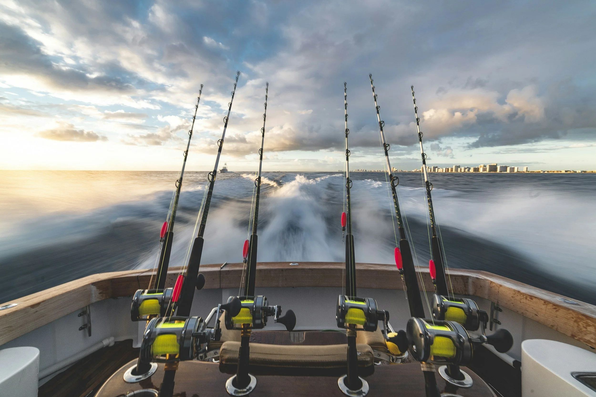 Multiple fishing rods mounted on the back of a boat, with a view of the ocean and a distant shoreline with buildings, under a partly cloudy sky during sunset.