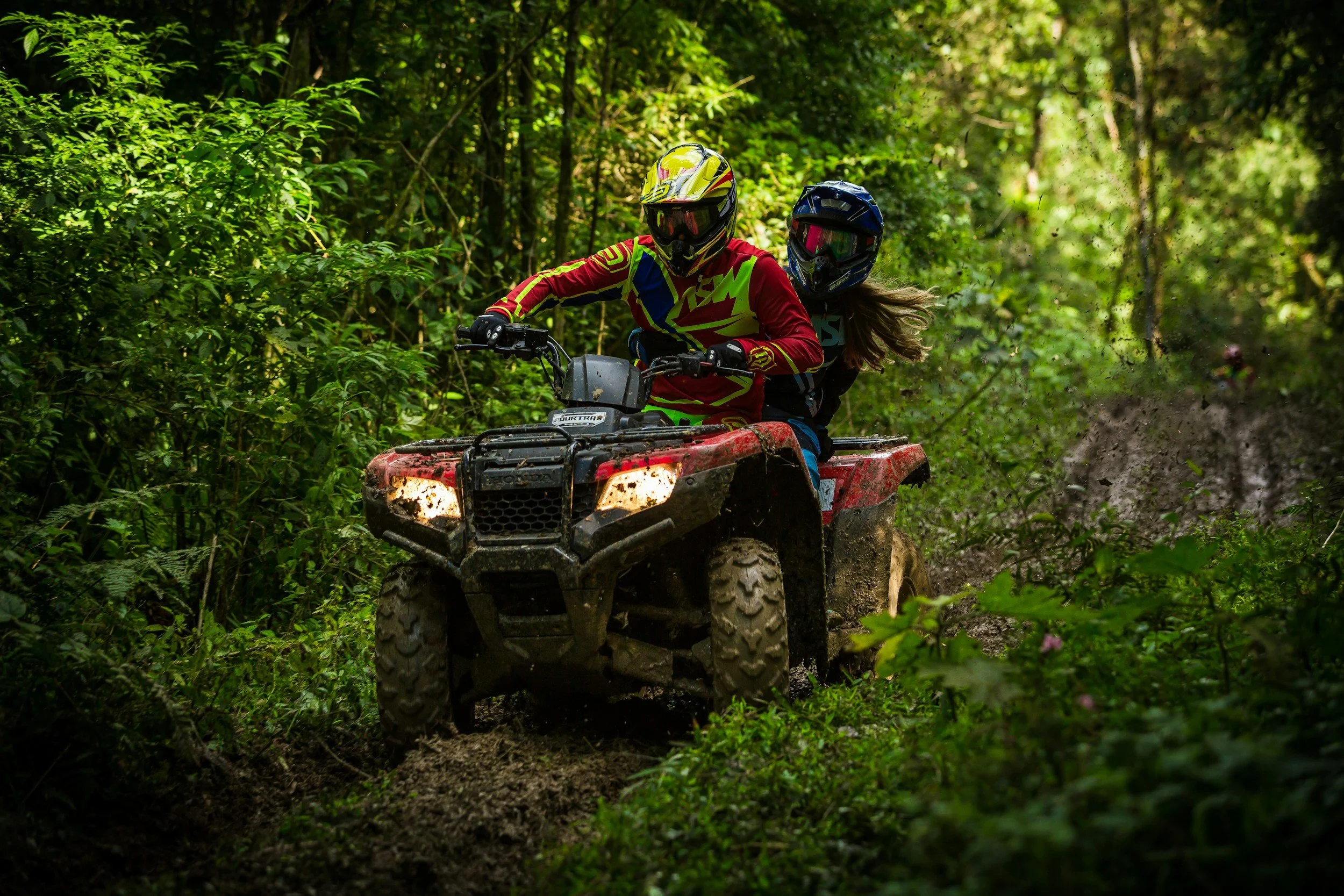 Two people riding an ATV through a forest trail, wearing helmets and colorful protective gear, with green foliage surrounding them.