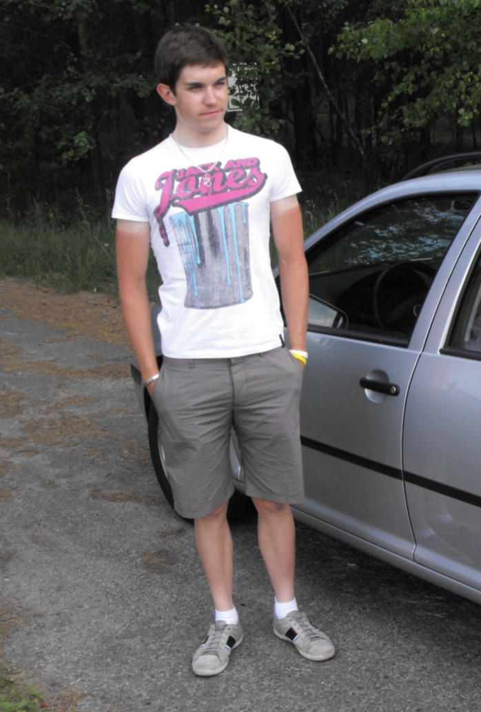 Junger Mann steht neben einem grauen Auto, trägt ein weißes T-Shirt mit buntem Print, graue Shorts, Turnschuhe und eine Halskette, vor einem grünen, bewaldeten Hintergrund.