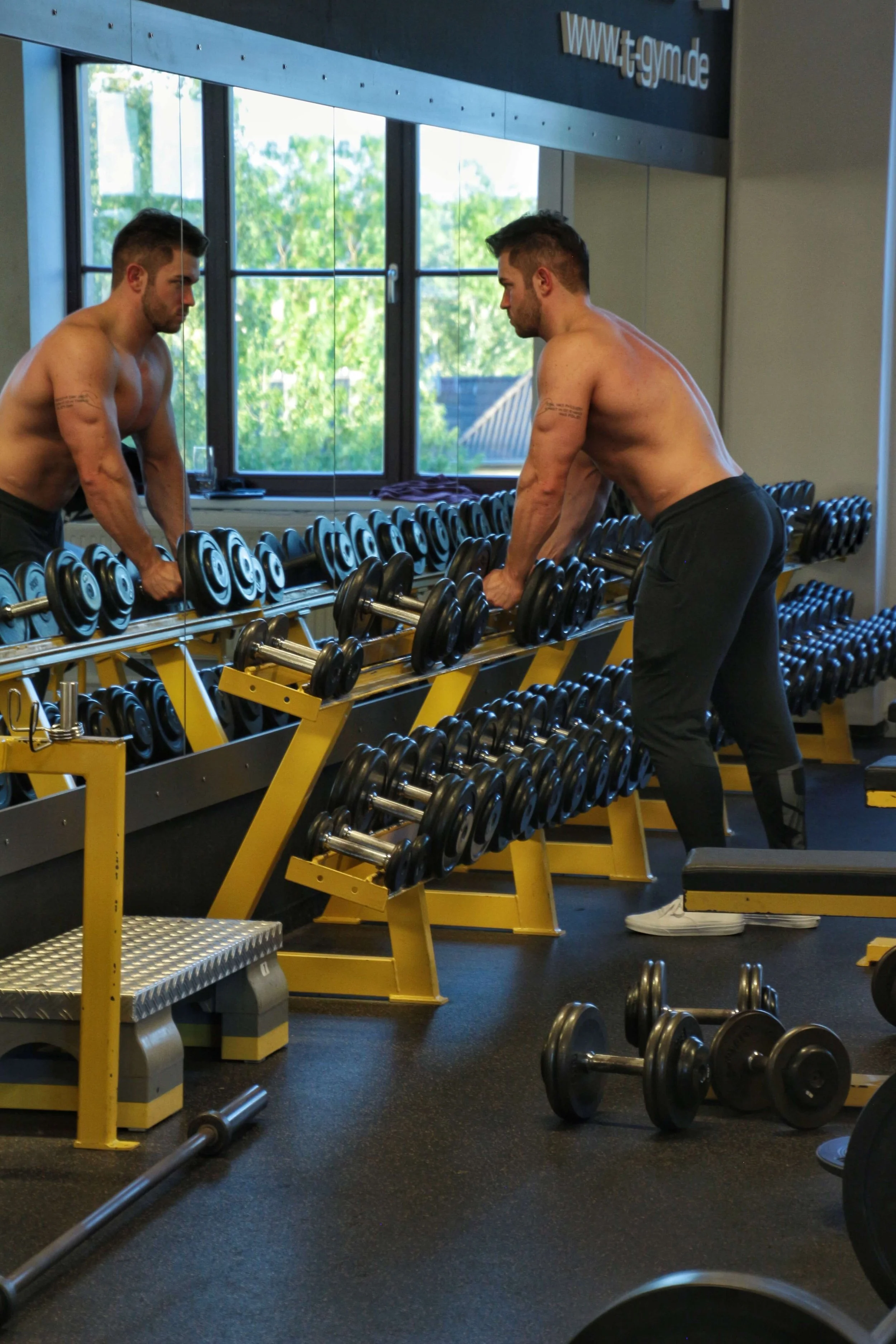 Ein athletischer Mann betrachtet mit entschlossener Miene sein Spiegelbild im Fitnessstudio und greift schwere Hanteln. Er bereitet sich auf ein intensives Training vor.