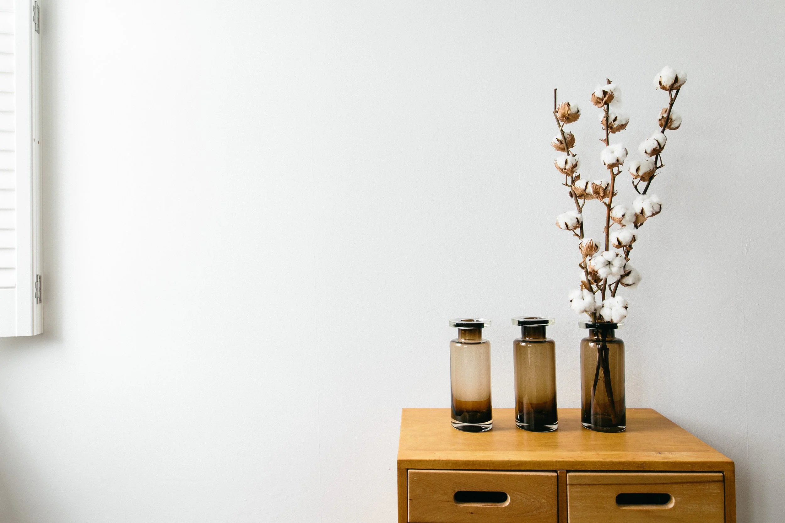Cotton stems in three brown glass vases on a wooden surface against a white wall.