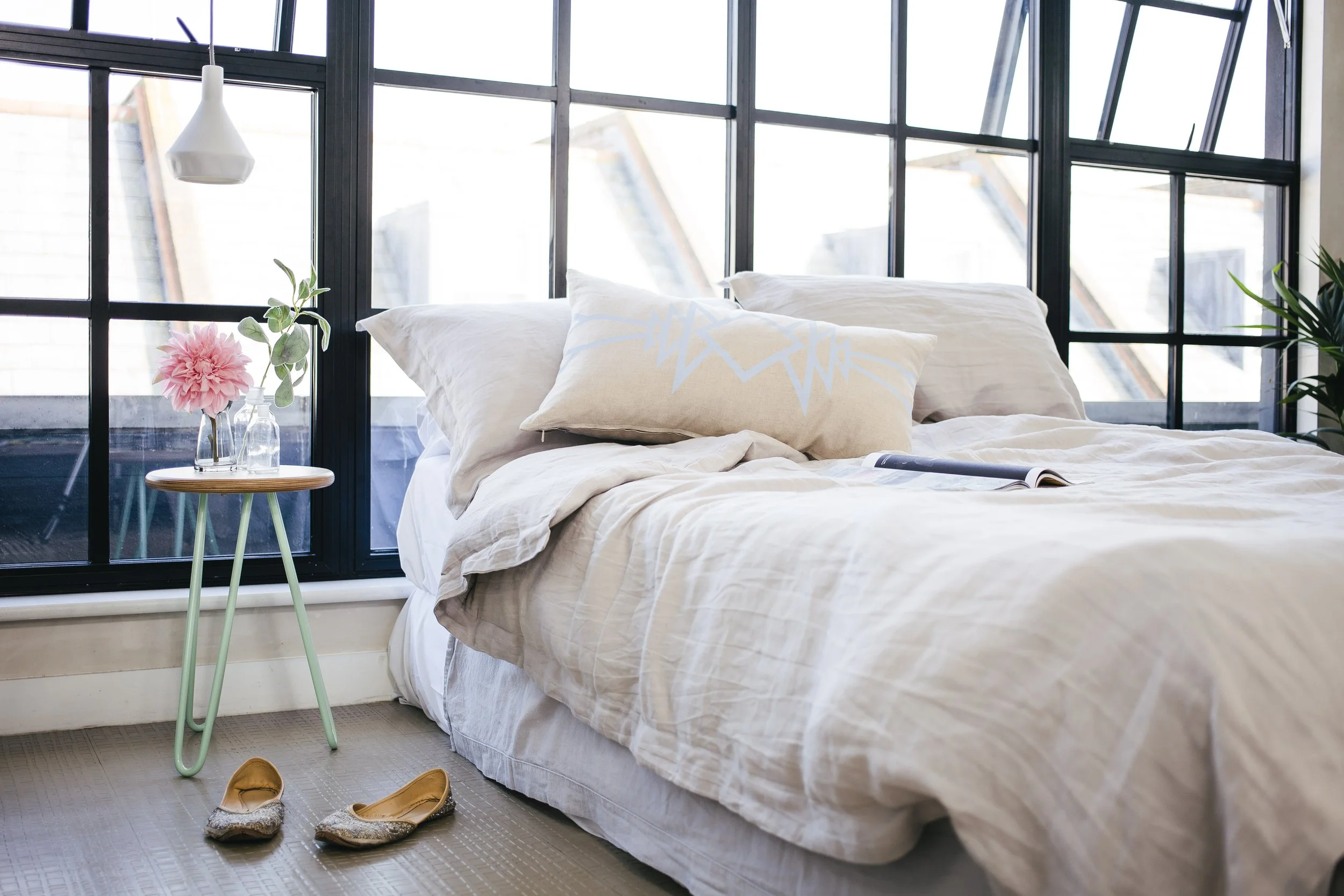 Light-filled bedroom styled with neutral linen bedding, a mint-green side table and pink flowers.