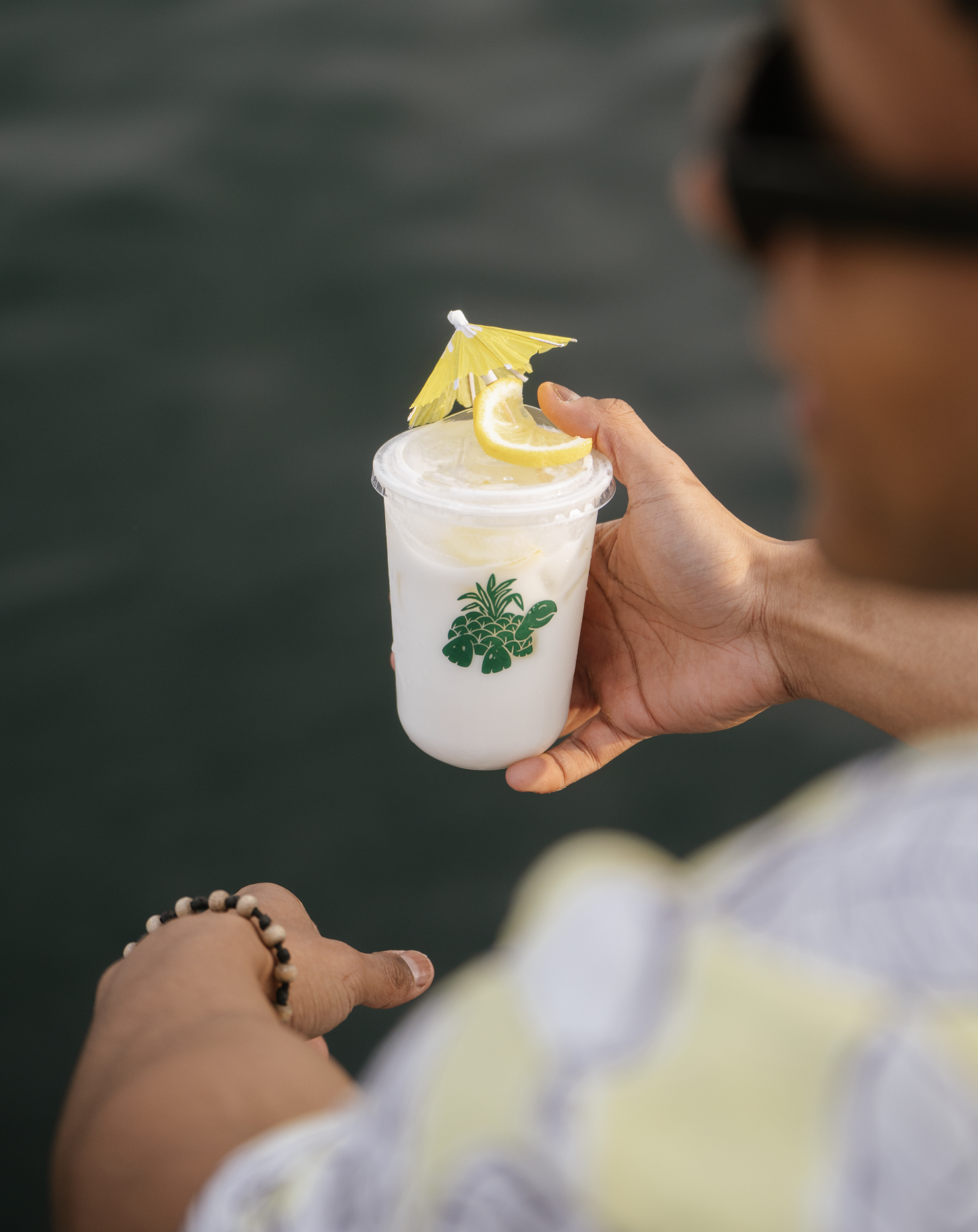 A person holding a drink with a lemon wedge and a yellow paper umbrella against a dark water background.