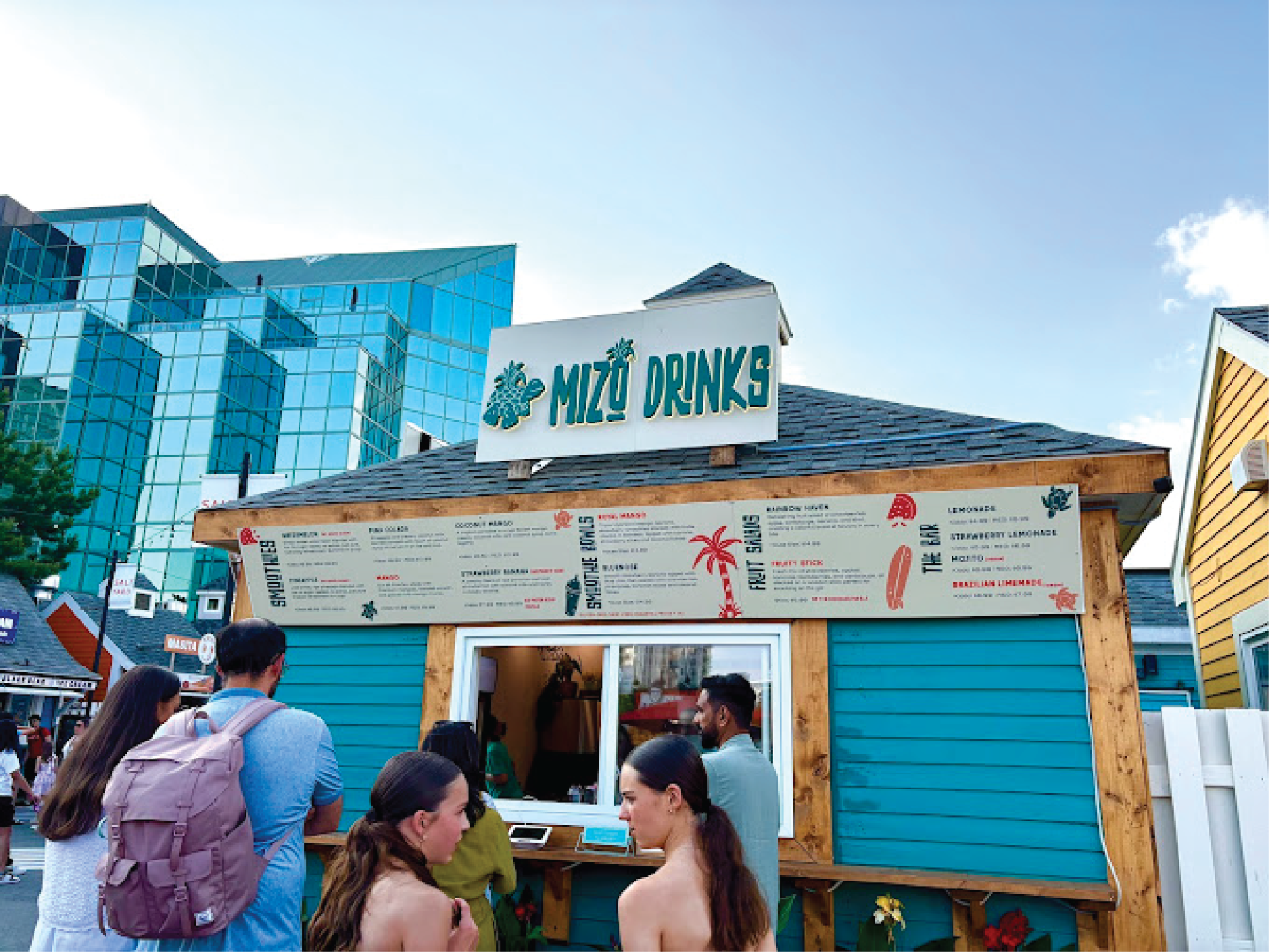 People standing in line outside a small blue food stand named 'Mizo Drinks' with a large menu board, located in an urban area with modern glass buildings in the background on a sunny day.