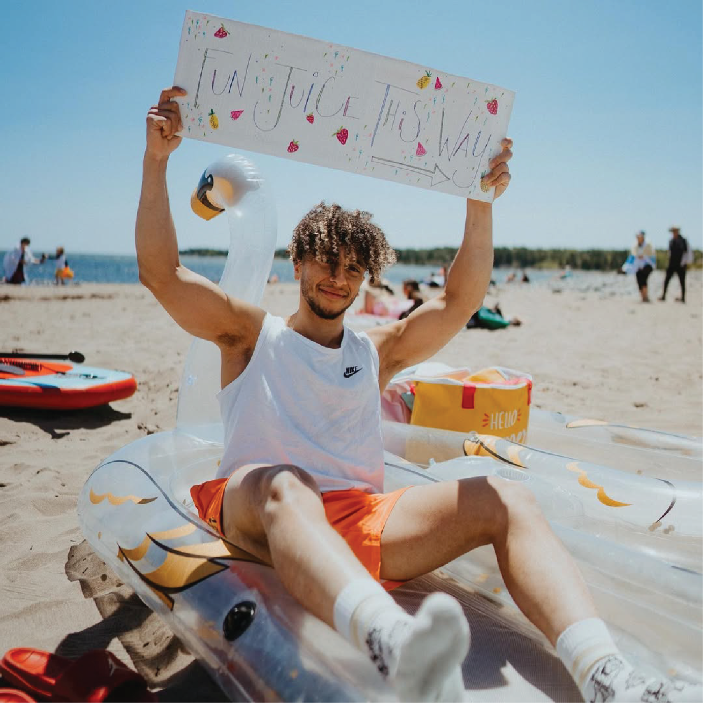 A young man sitting on a beach inflatable float, holding a colorful sign that says 'Fun, Juice, This Way' while smiling at the camera. There are people in the background near the water, a paddleboard on the sand, and a bright, sunny sky.
