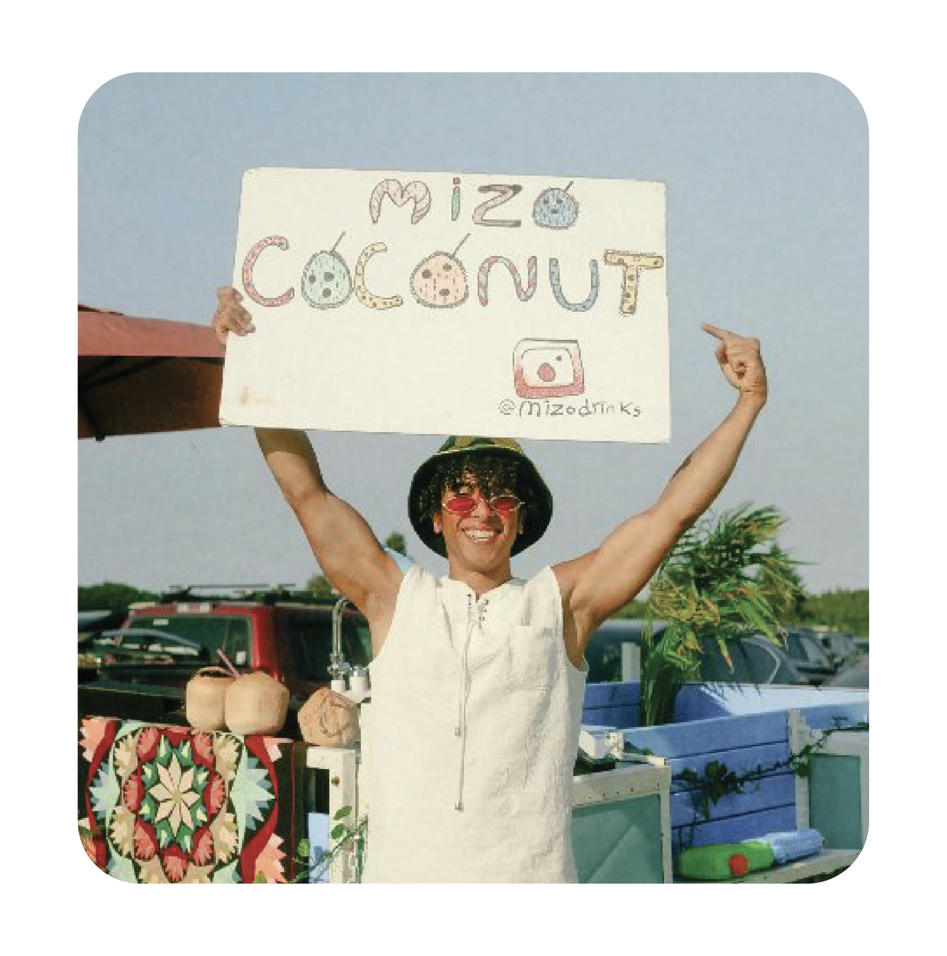 A smiling woman wears sunglasses and a wide-brimmed hat, holding a sign that says 'Mi zoo coconut' with a plush coconut and a crochet coconuts on it, outdoors near palm trees and parked cars.