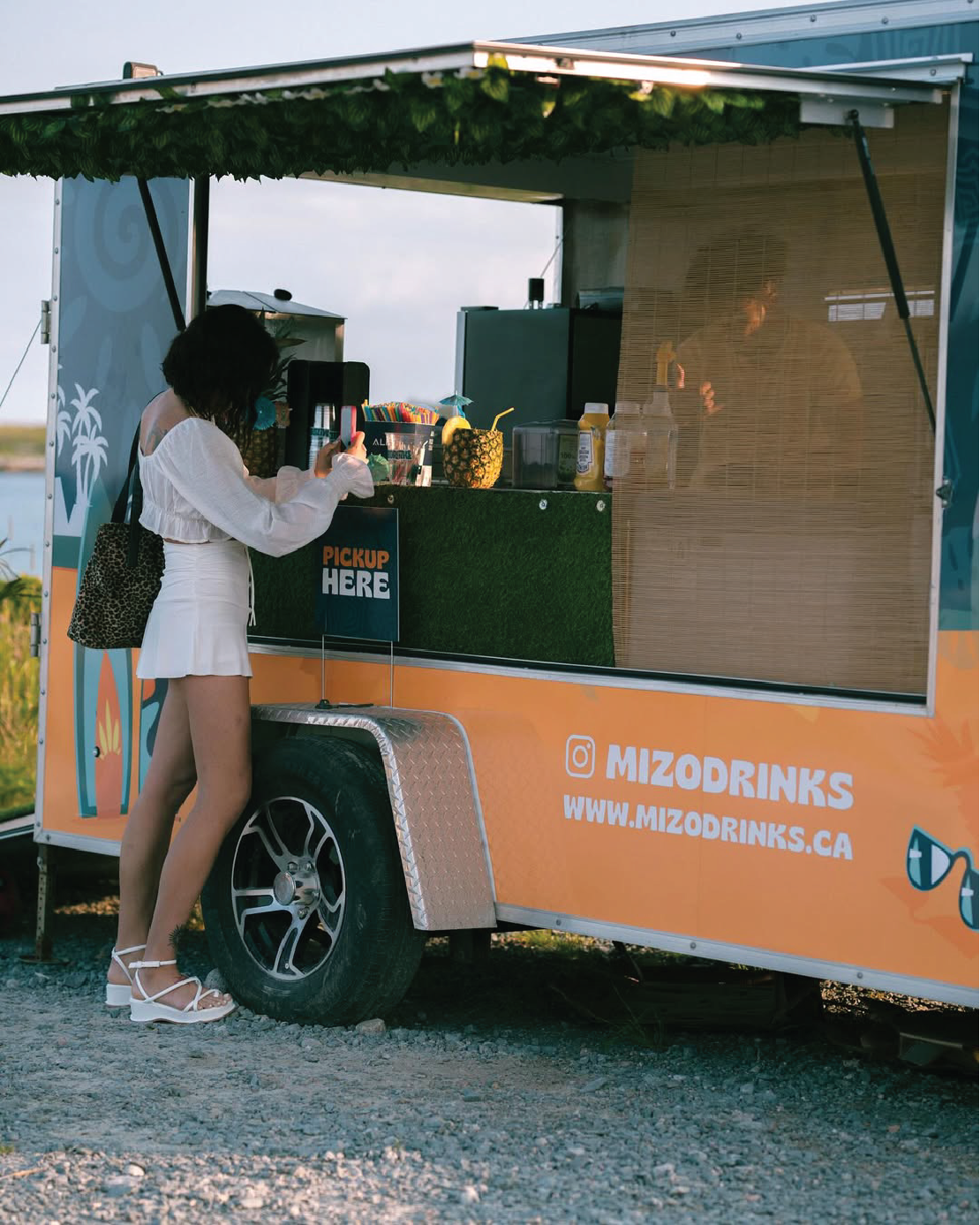 A woman in white clothing and sandals standing at a colorful drink truck, taking a photo with her phone. The truck has a sign that reads 'PICKUP HERE' and the website www.mizodrinks.ca, with some drinks and a pineapple decoration visible on the counter.