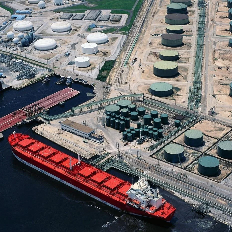 Immagine di un impianto industriale con serbatoi di varie dimensioni, strutture e un grande cargo rosso ancorato al porto.