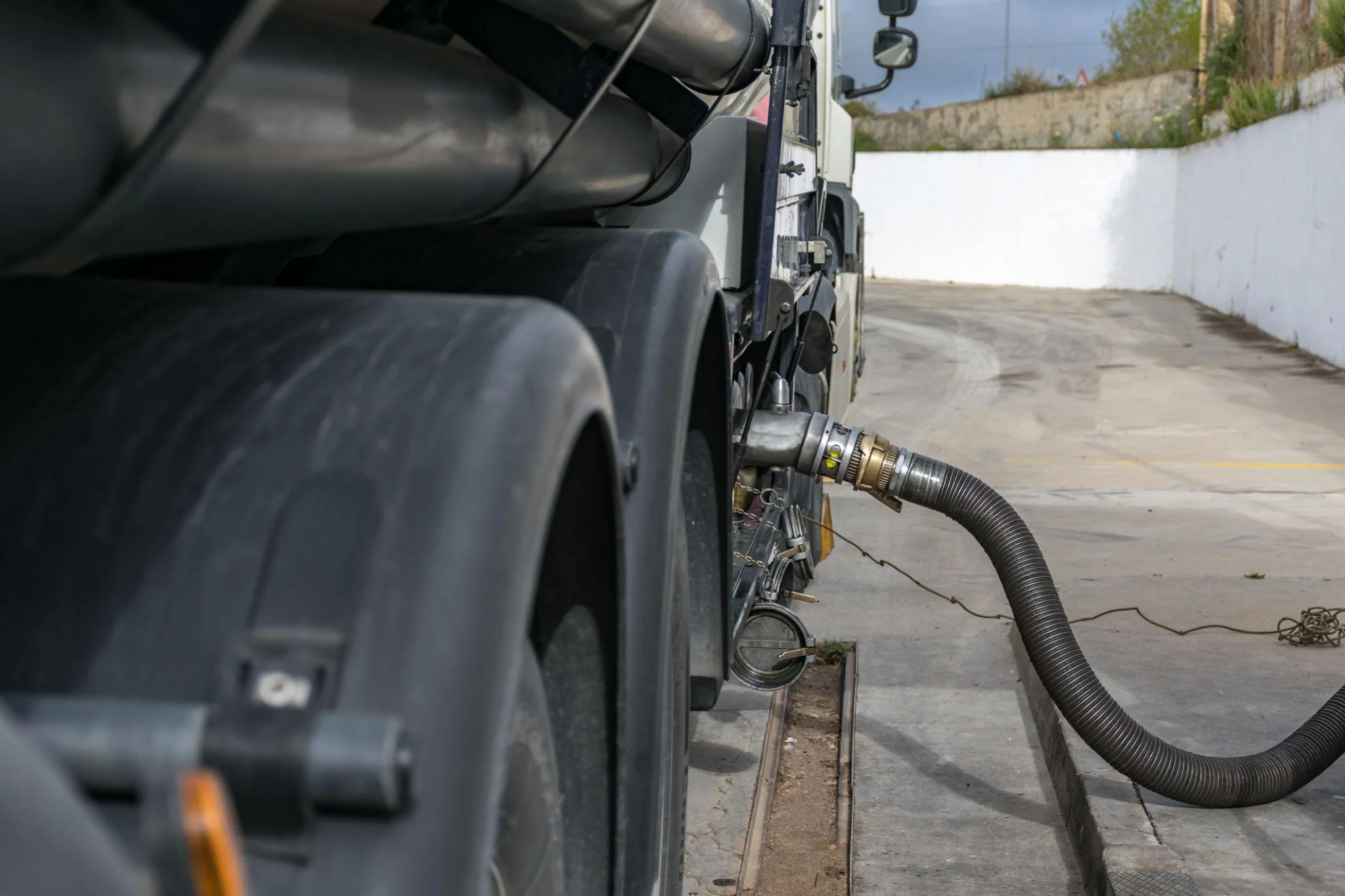Un teaser di un camion di acqua con un tubo collegato alla parte posteriore, presso un'area all'aperto con pareti di recinzione e un muro bianco.