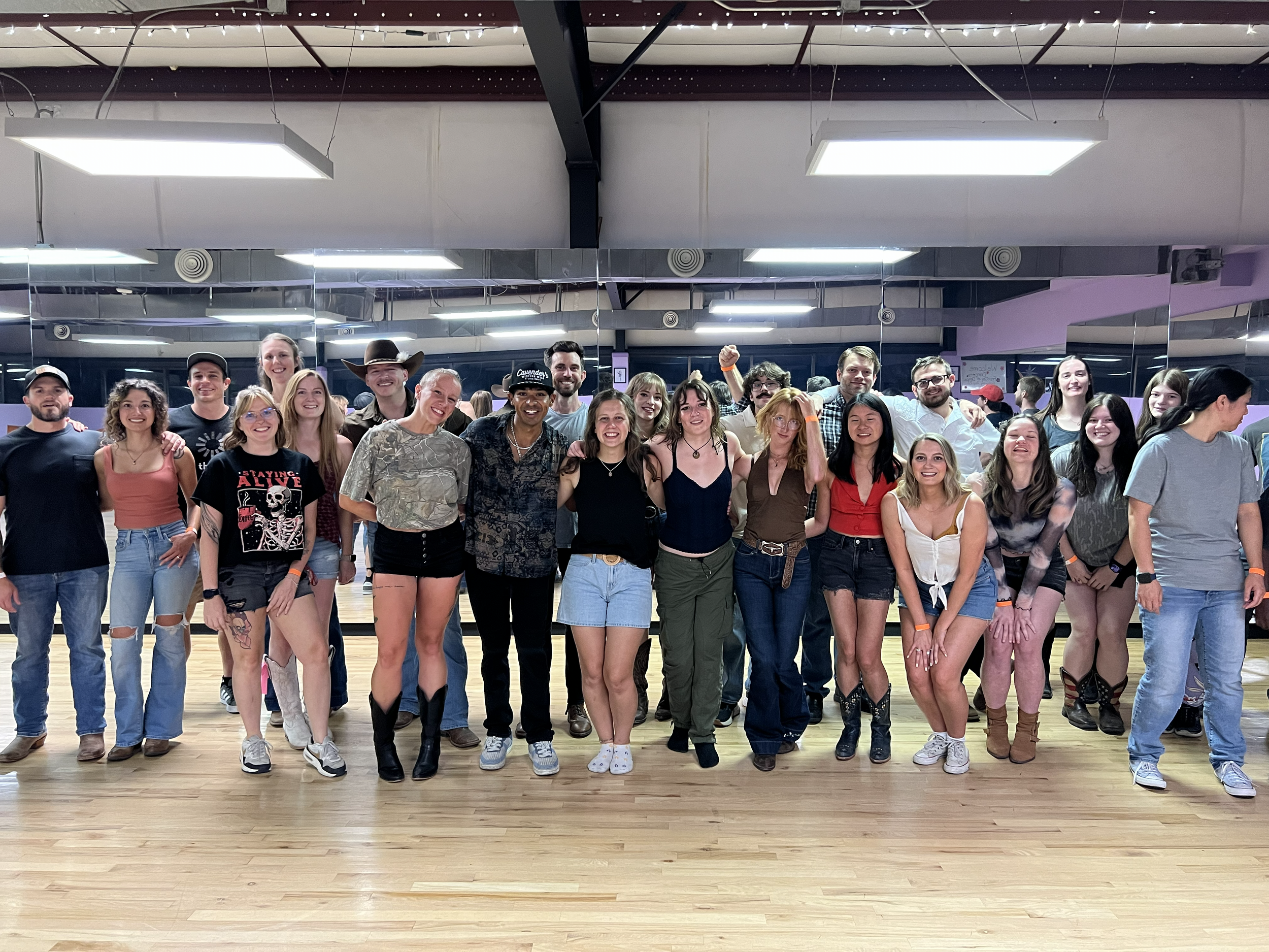 Large group posing for a photo in a mirrored dance studio with wood floors and overhead lights