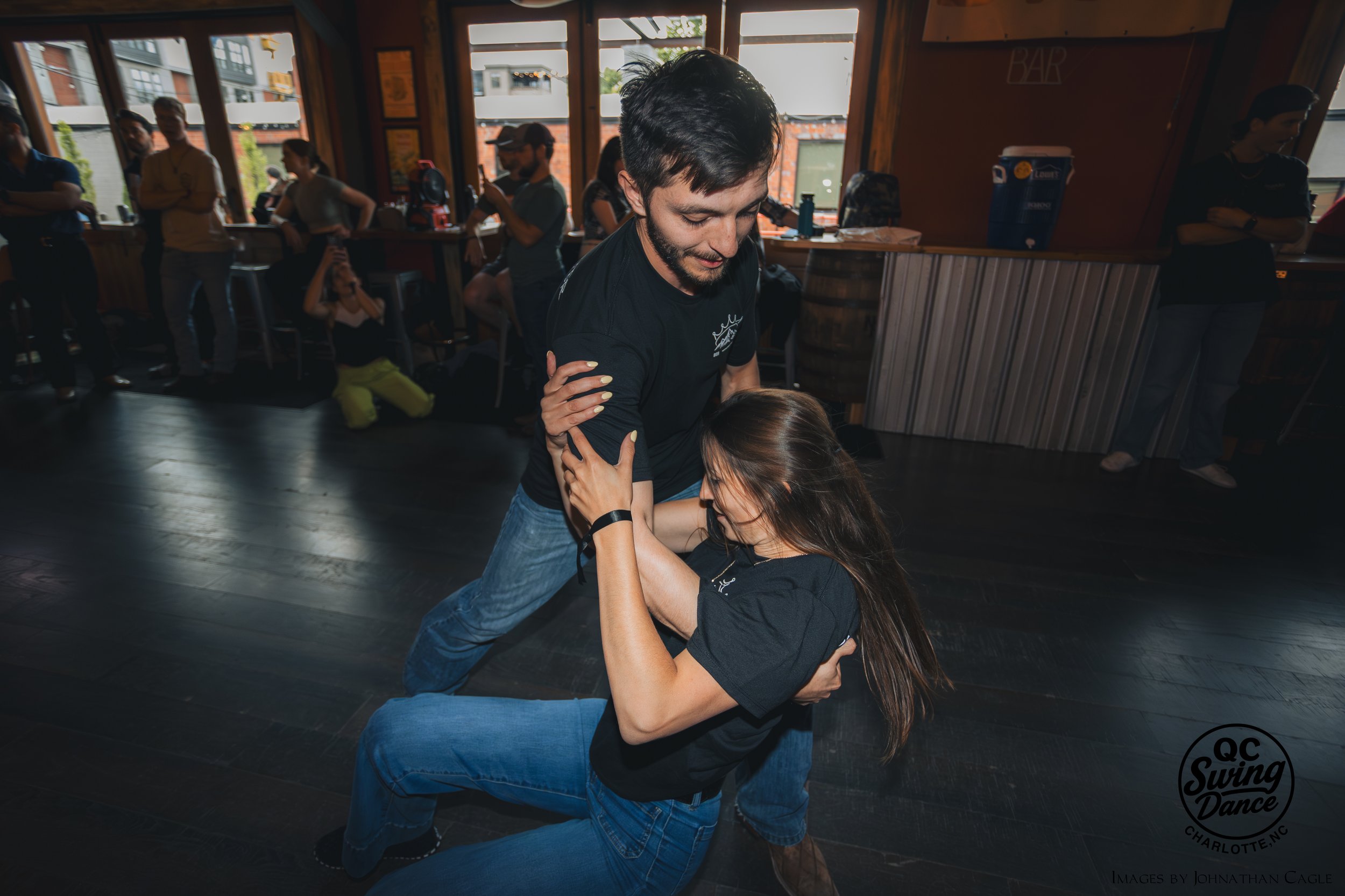A man and woman are dancing together in a lively indoor setting with other people in the background. The man is holding the woman around her waist, and the woman is holding his shoulder. They appear to be enjoying their dance.