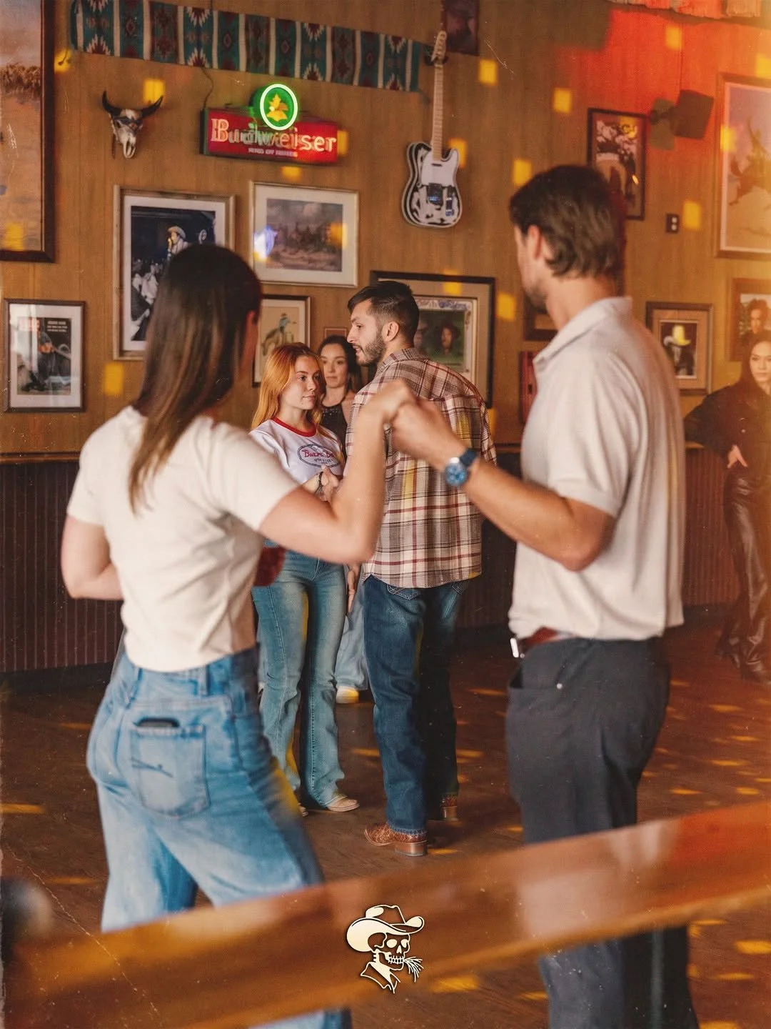 People dancing and socializing in a bar with wooden walls decorated with framed pictures, a guitar, and a neon Budweiser sign.