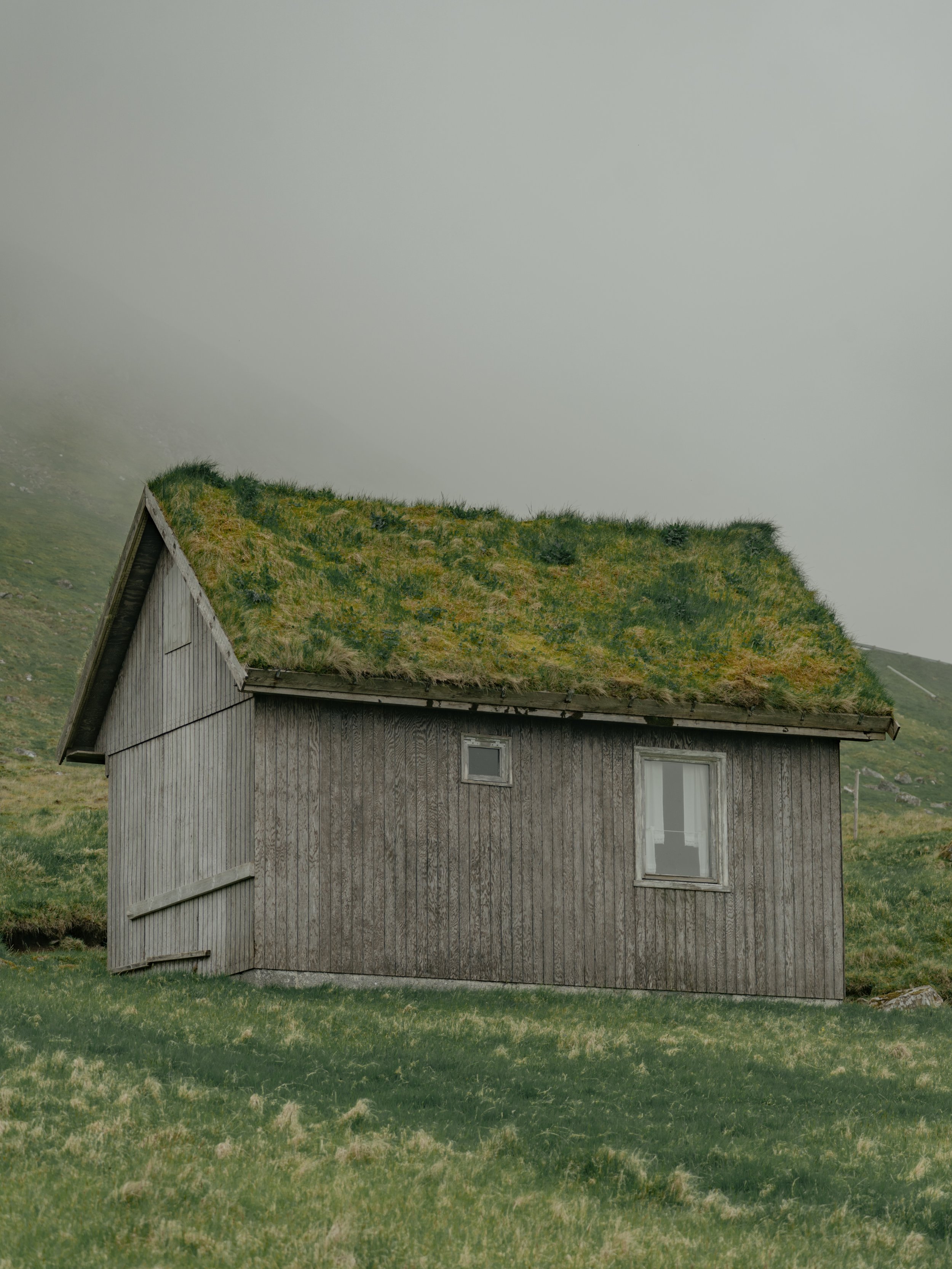 A house with wooden siding and a grass-covered roof in a rural, mountainous area with foggy sky.