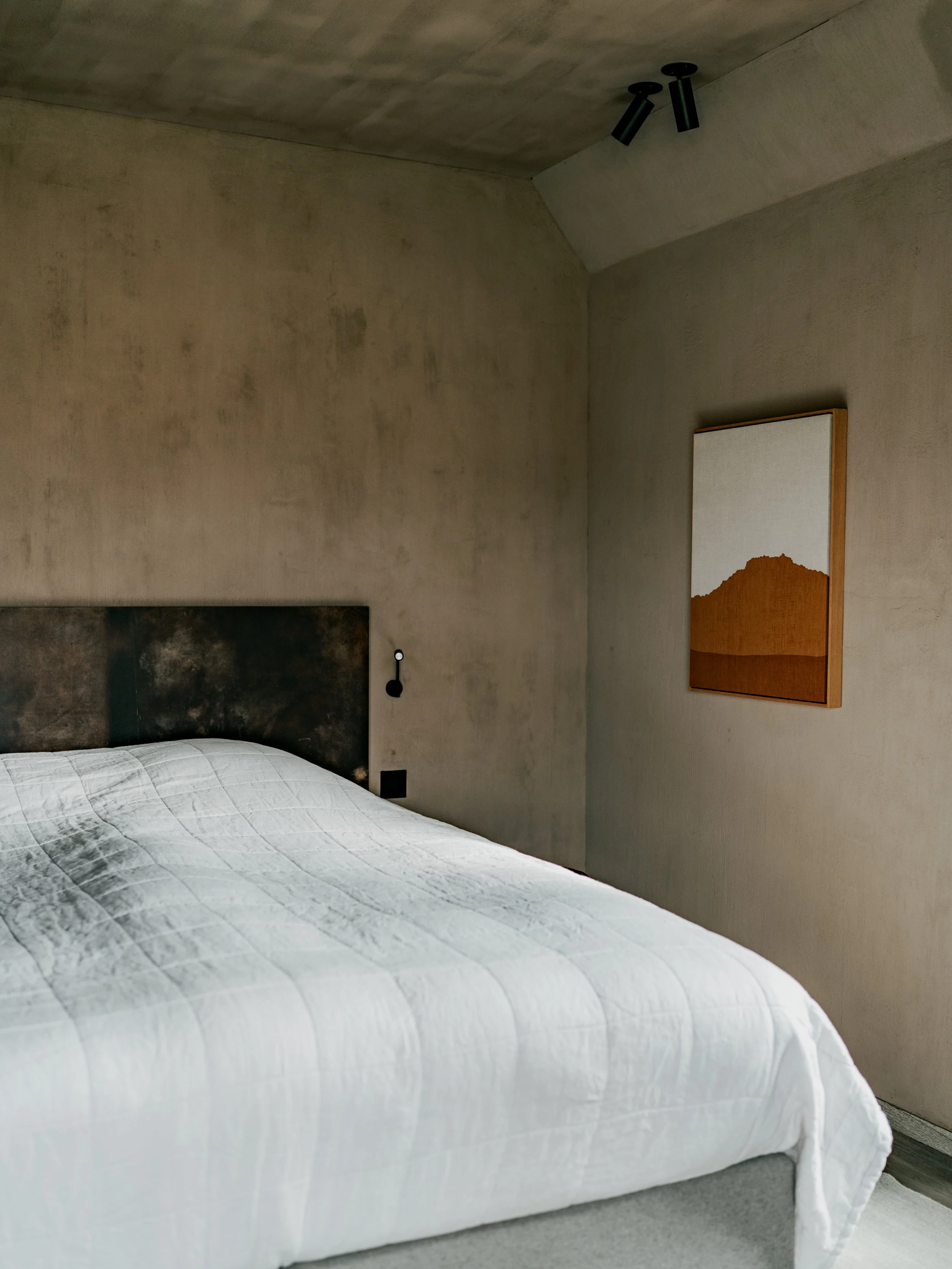 A minimalist bedroom with a bed covered in a white quilt, a textured beige wall, and a modern wall art piece with a mountain landscape and orange tones, with ceiling-mounted black light fixtures.