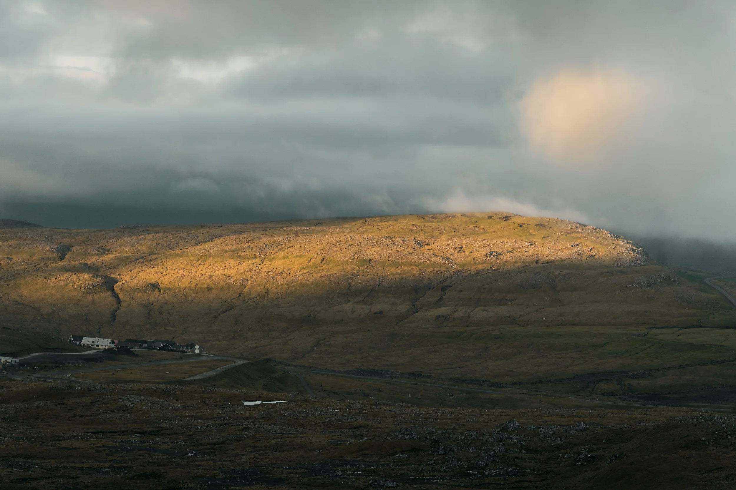 A landscape with a cloudy sky, hilly terrain, and a small settlement of houses at the bottom.