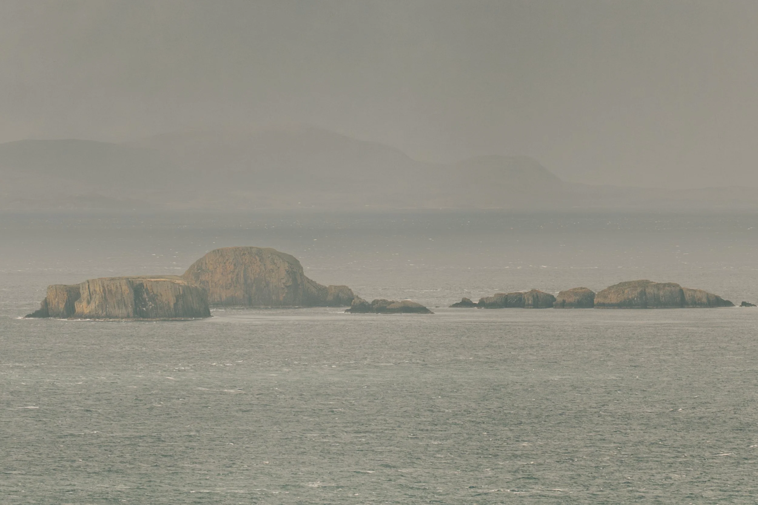 Overcast ocean view with large rocks in the water and distant mountains on the horizon.