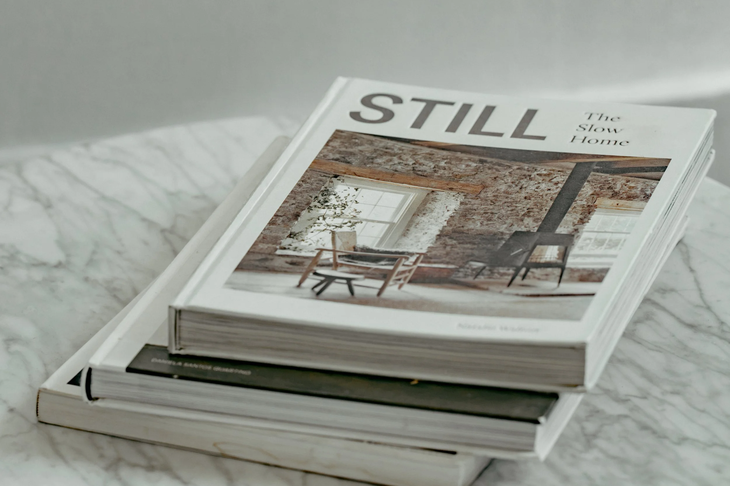 Stack of magazines with the top magazine titled 'The Still Home' featuring an interior design photograph on the cover, placed on a white marble surface.
