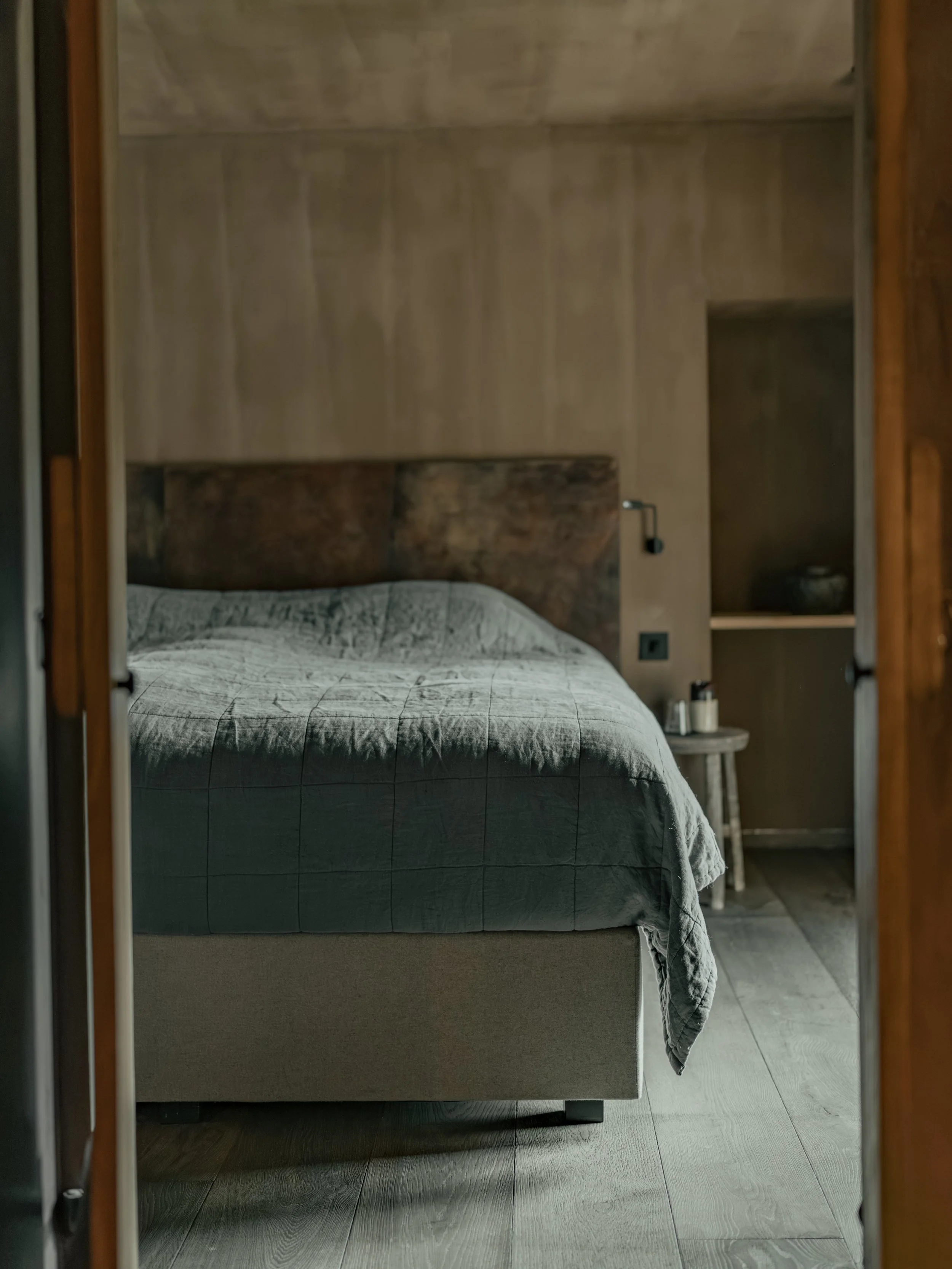 A bedroom seen through a doorway, featuring a bed with a textured gray quilt, wooden headboard, side table with decor, and a built-in shelf with a decorative object, in a modern, minimalistic style.