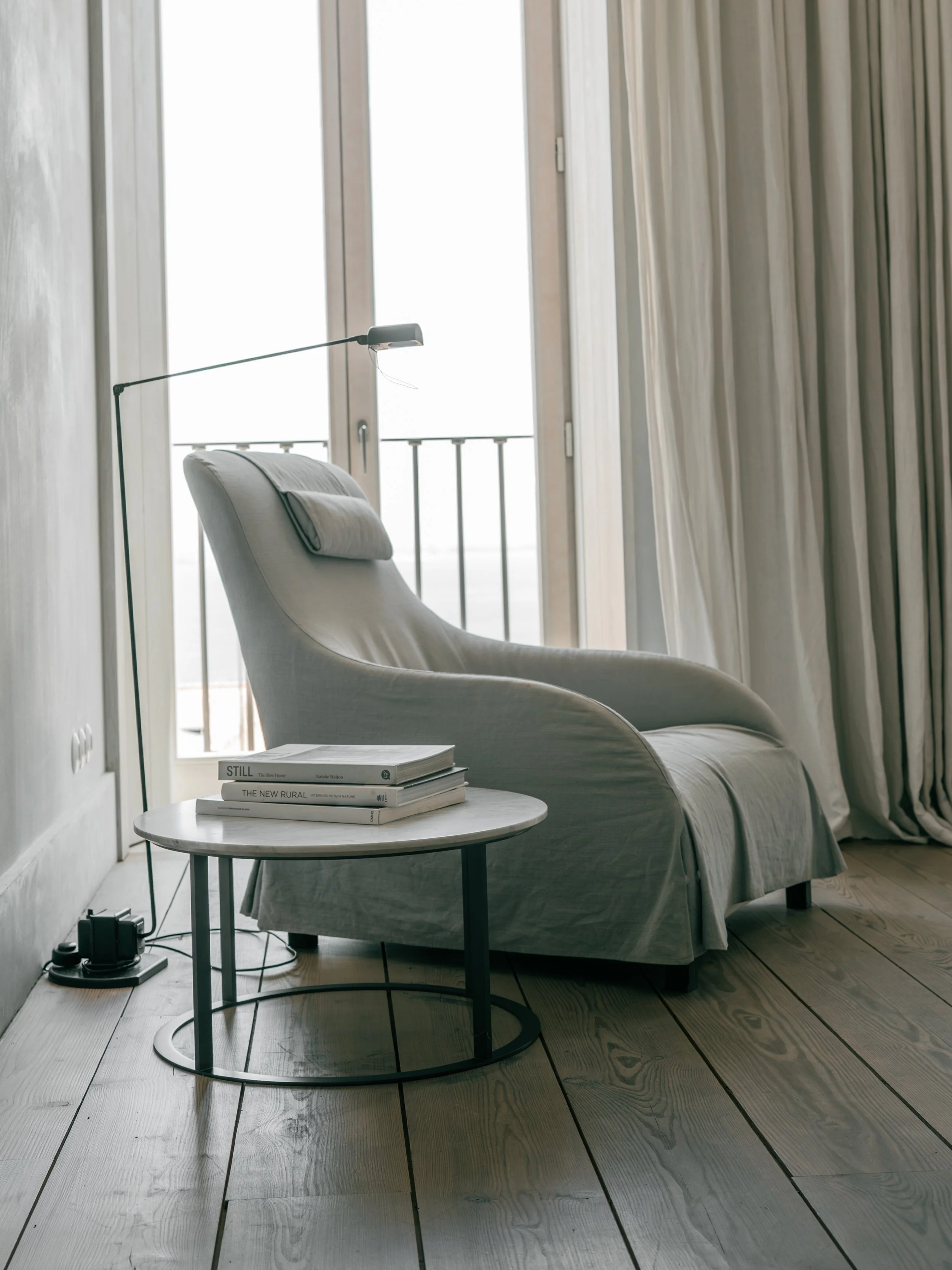 A cozy corner with a beige armchair with a pillow, a round white table with books, a floor lamp, near large sliding glass doors with curtains, and a balcony with a railing outside.