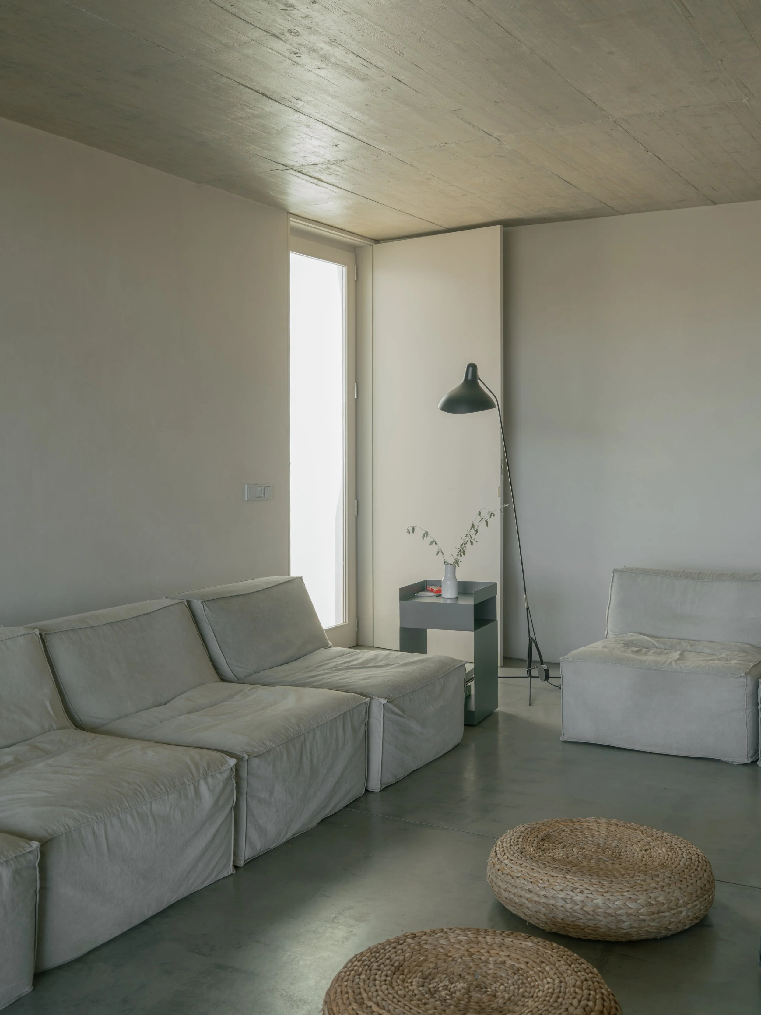 Living room with white sofas, a gray side table with a white vase and plant, black floor lamp, white walls, a door, and woven poufs on a concrete floor.