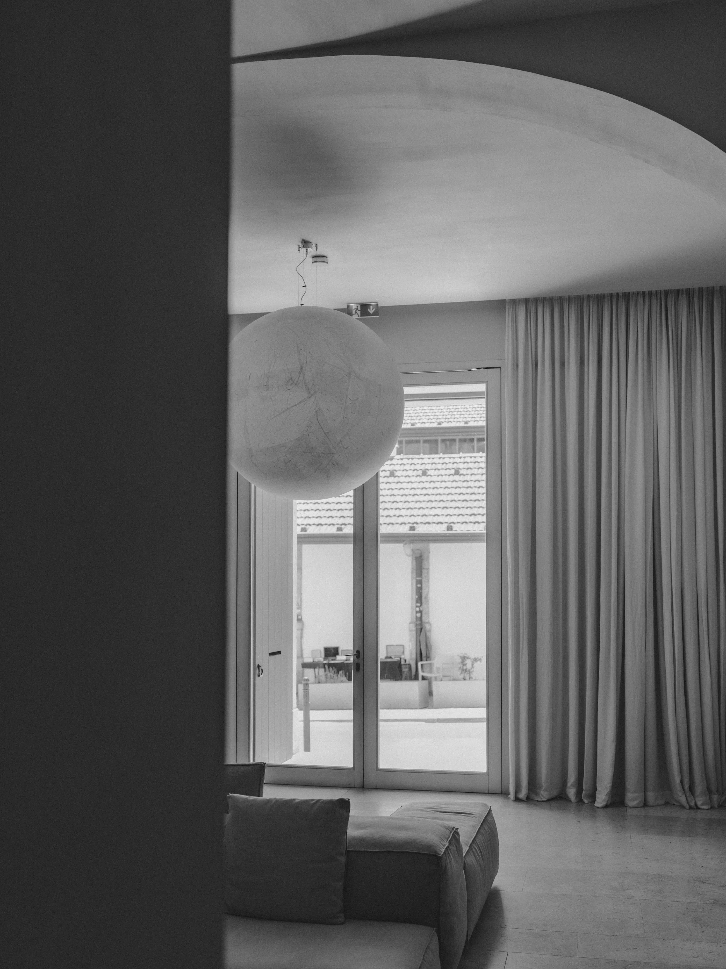 A black and white photo of a living room with a large window and curtains. There's a couch with cushions, a spherical pendant light hanging from the ceiling, and an exit sign above the door.