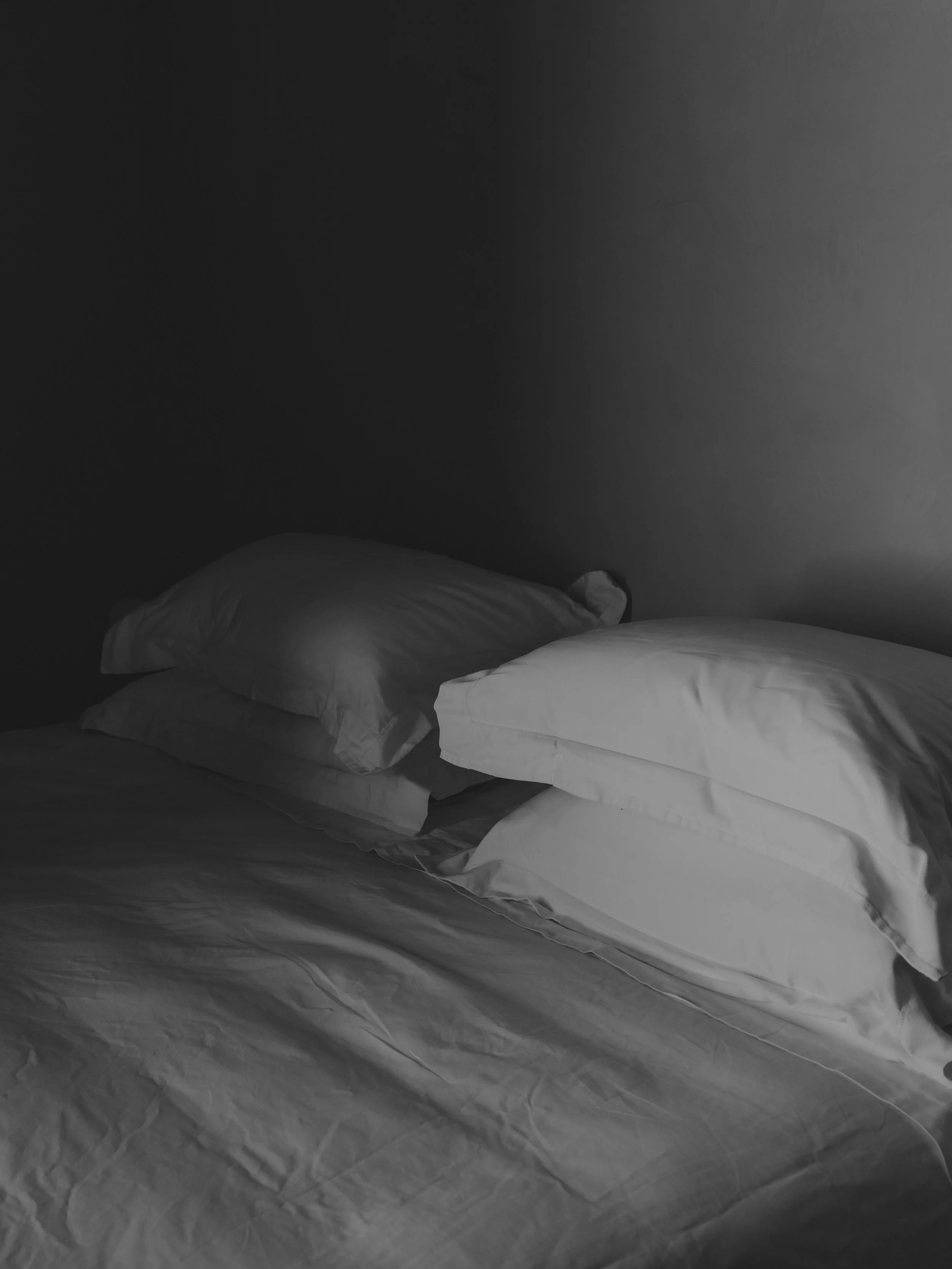 A bed with white sheets and pillows in a dimly lit room with a plain wall.