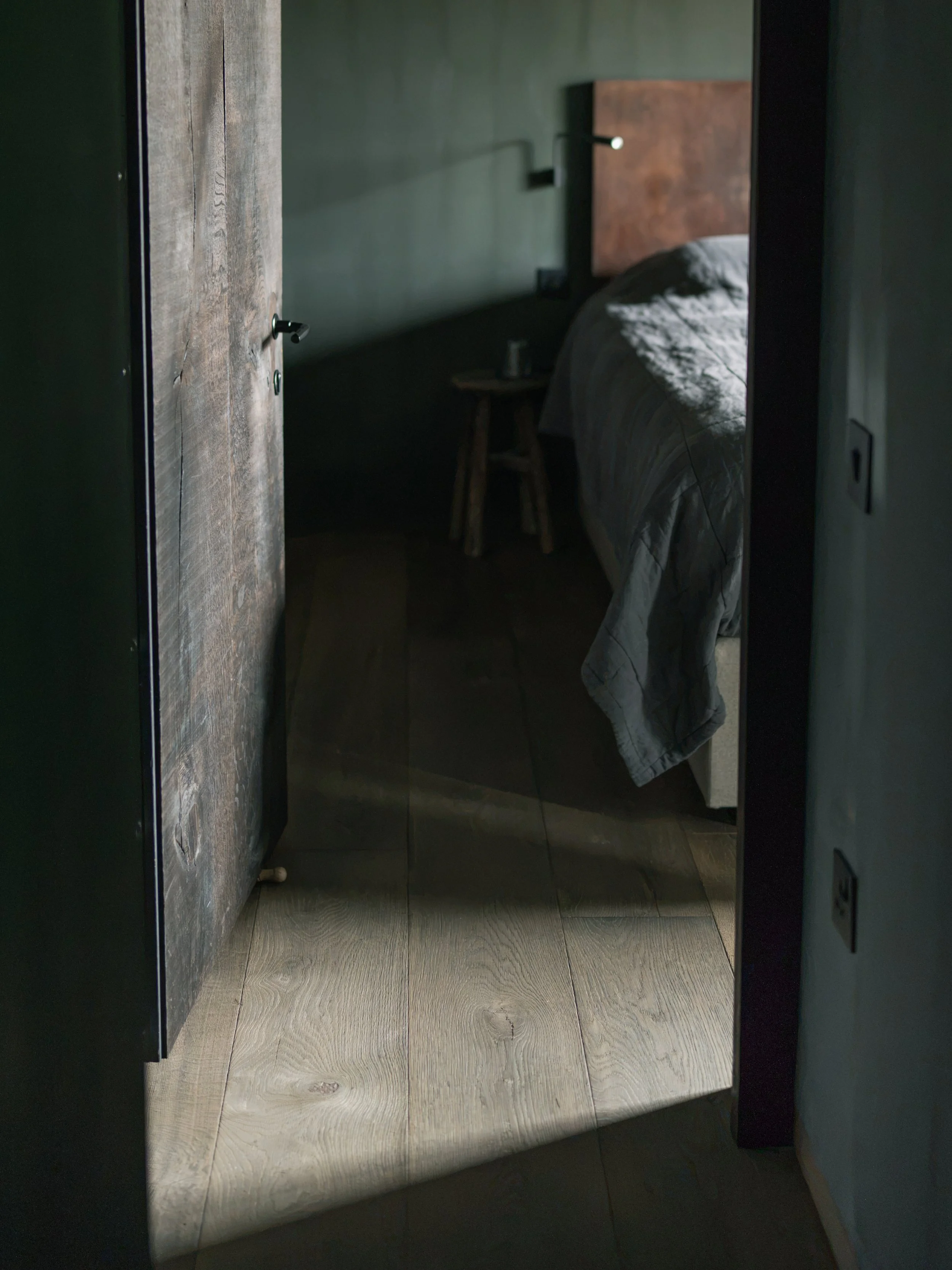 A reflection of a wooden bed with a dark headboard, a small round wooden side table with a cup, and a wall-mounted light in a dimly lit room, as seen through a mirror on the wall.