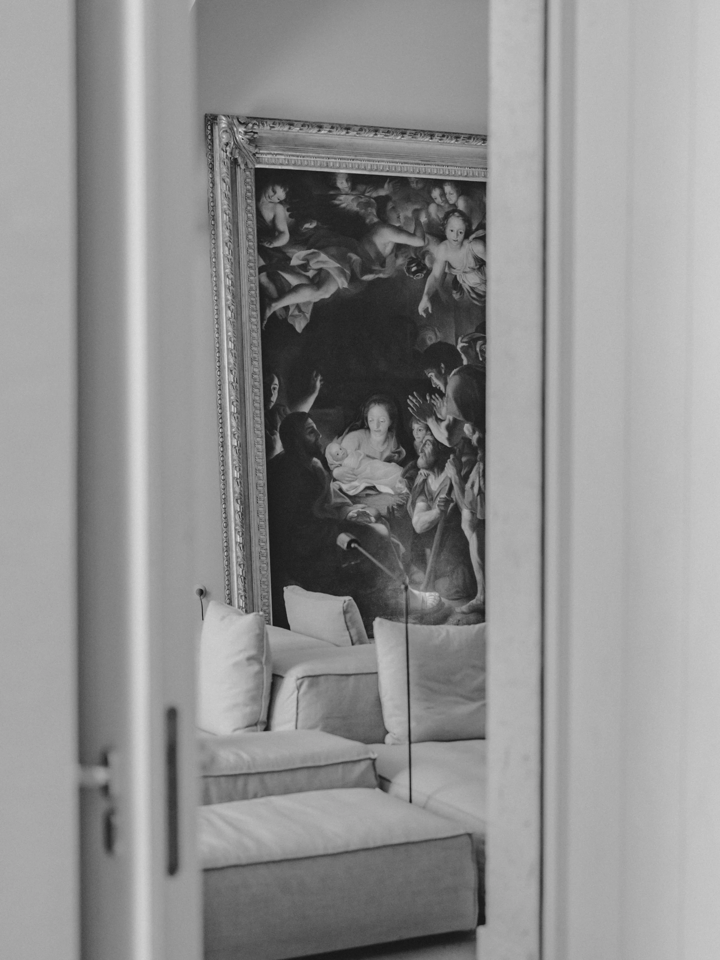A reflection in a mirror showing a living room with a framed religious painting, a sofa with pillows, and the edge of a door frame.
