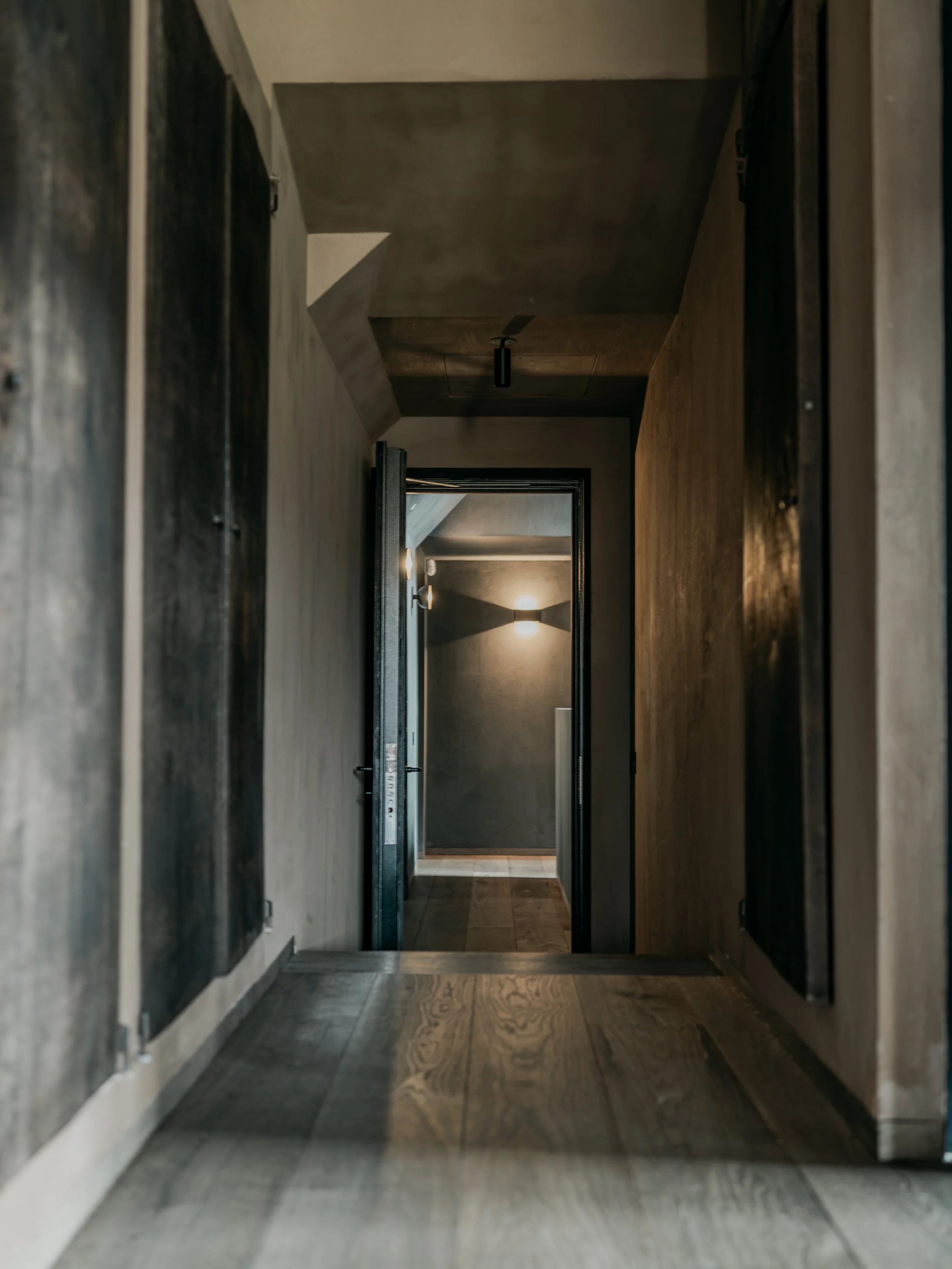 A narrow hallway with dark wood floors and walls, leading to a lit room with a ceiling light fixture visible.