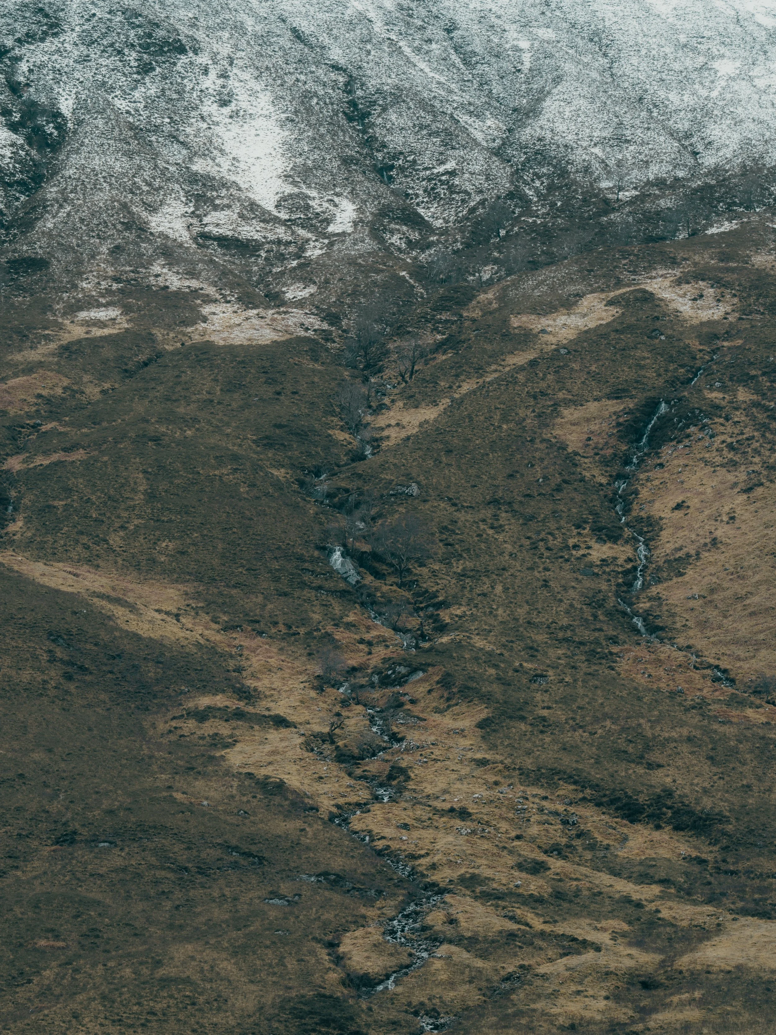 A mountain landscape with some snow on the upper slopes and a small stream running through the valley.