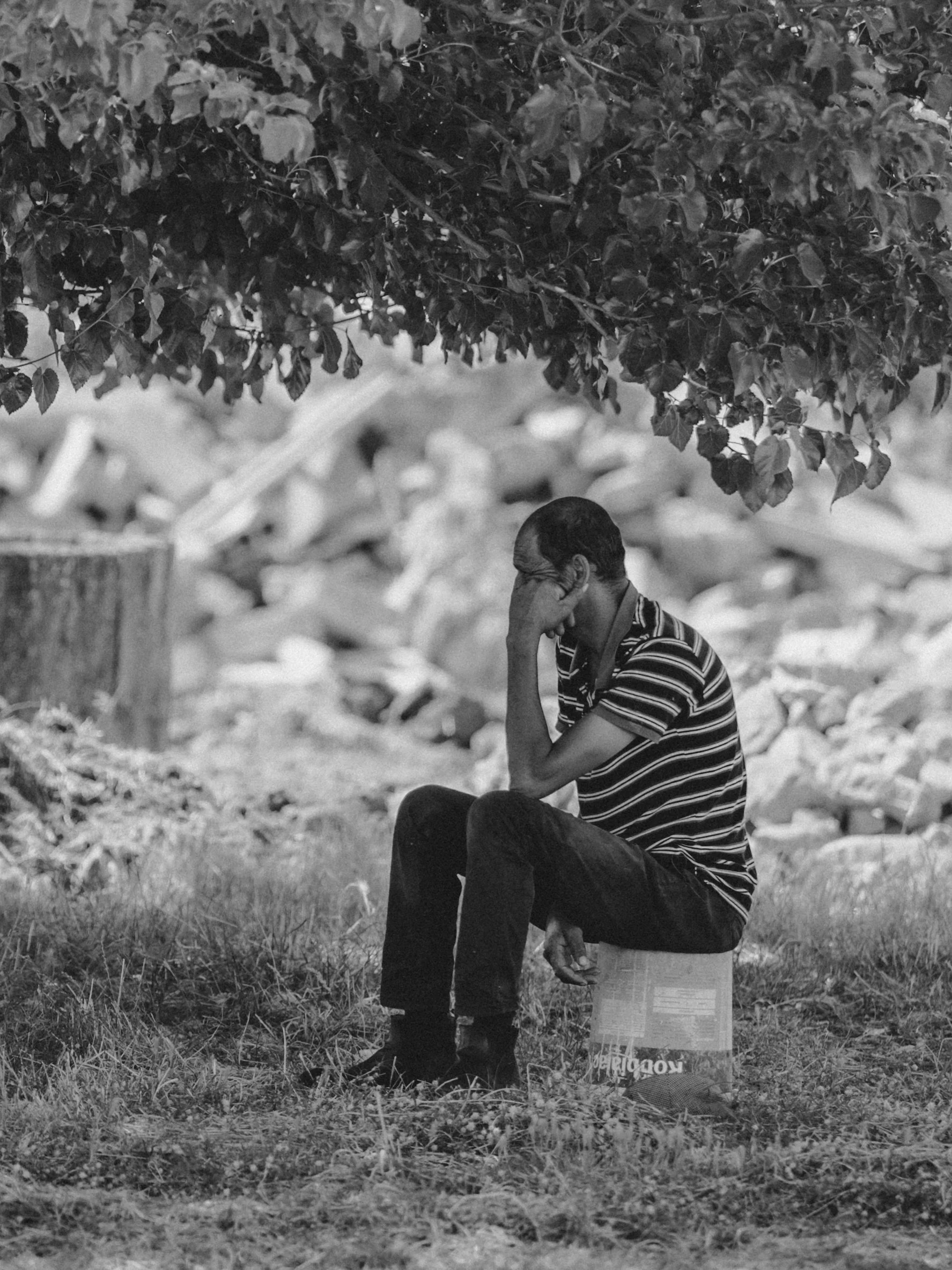 A man sitting on the ground under a tree, appearing to be deep in thought, with his hand covering his face, in a black and white photo.