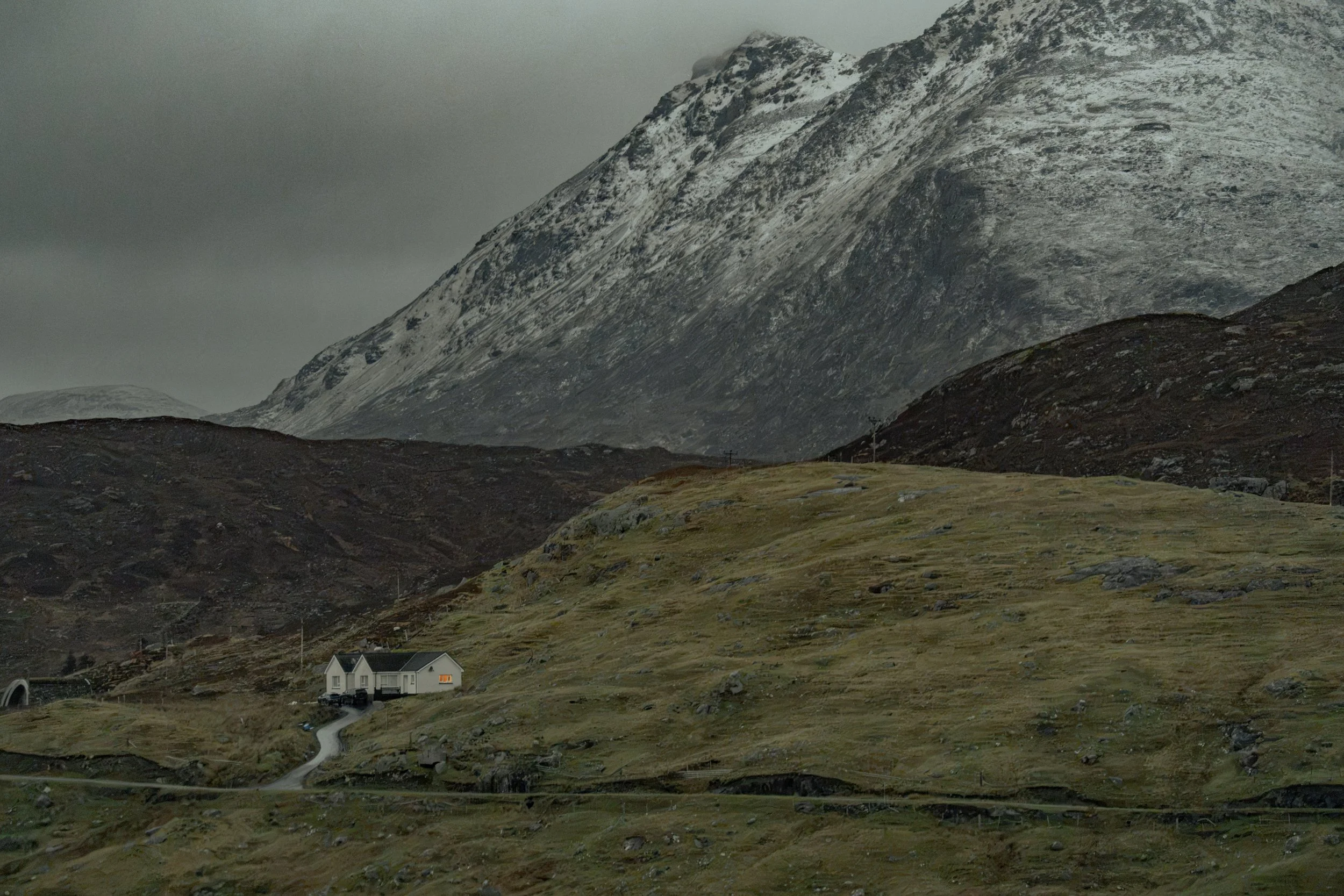 Small white house with dark roof in a hilly landscape, mountains with patches of snow in the background, cloudy sky.