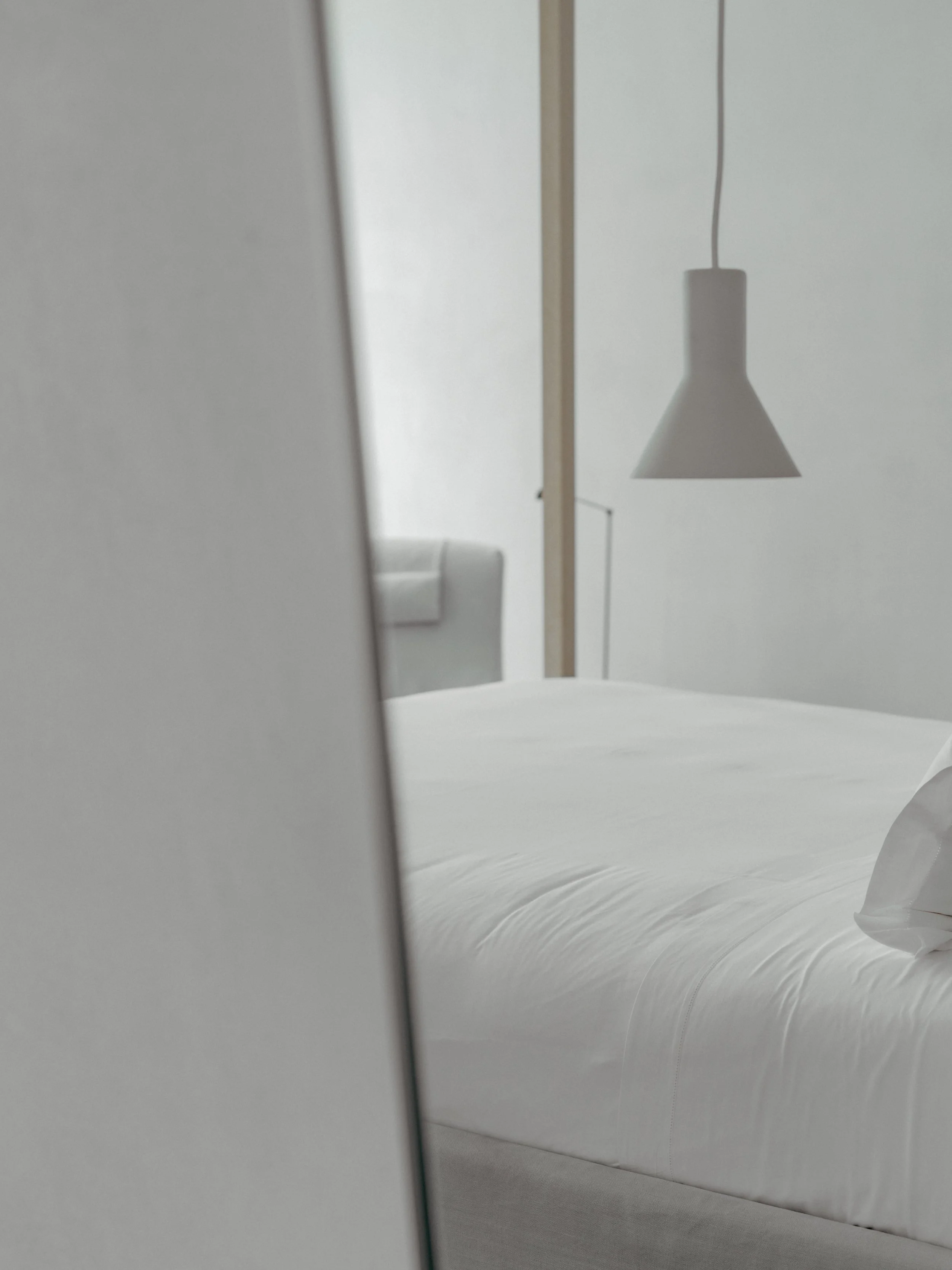 A bed with a white headboard and white bedsheets in a room with minimal decor. A modern, gray pendant light hangs from the ceiling.