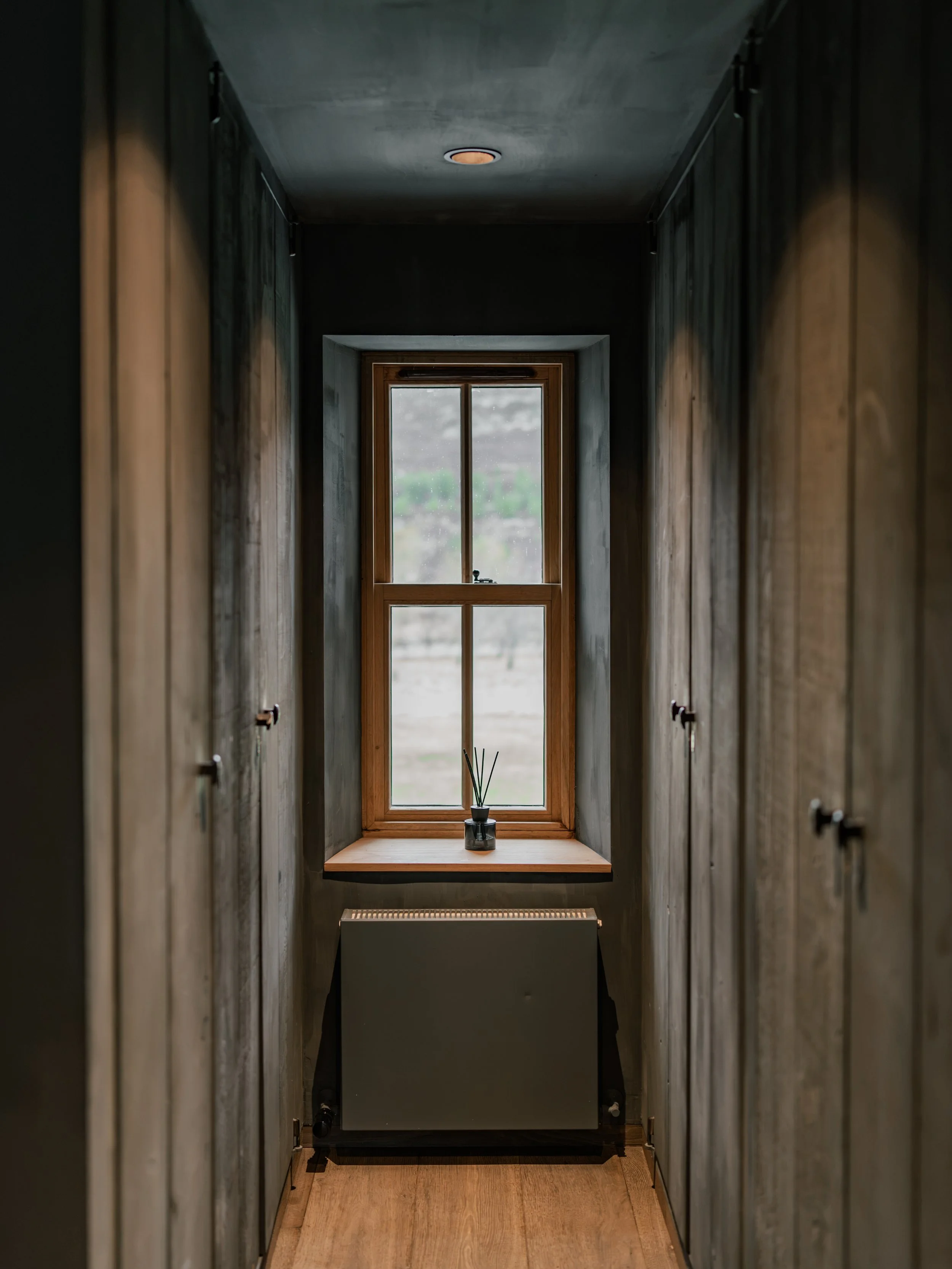 A narrow hallway with wooden floors and grey walls, featuring multiple small cabinets on either side. At the end of the hallway, a window with a wooden frame allows natural light inside and shows an outdoor scene with cloudy weather and greenery. A s