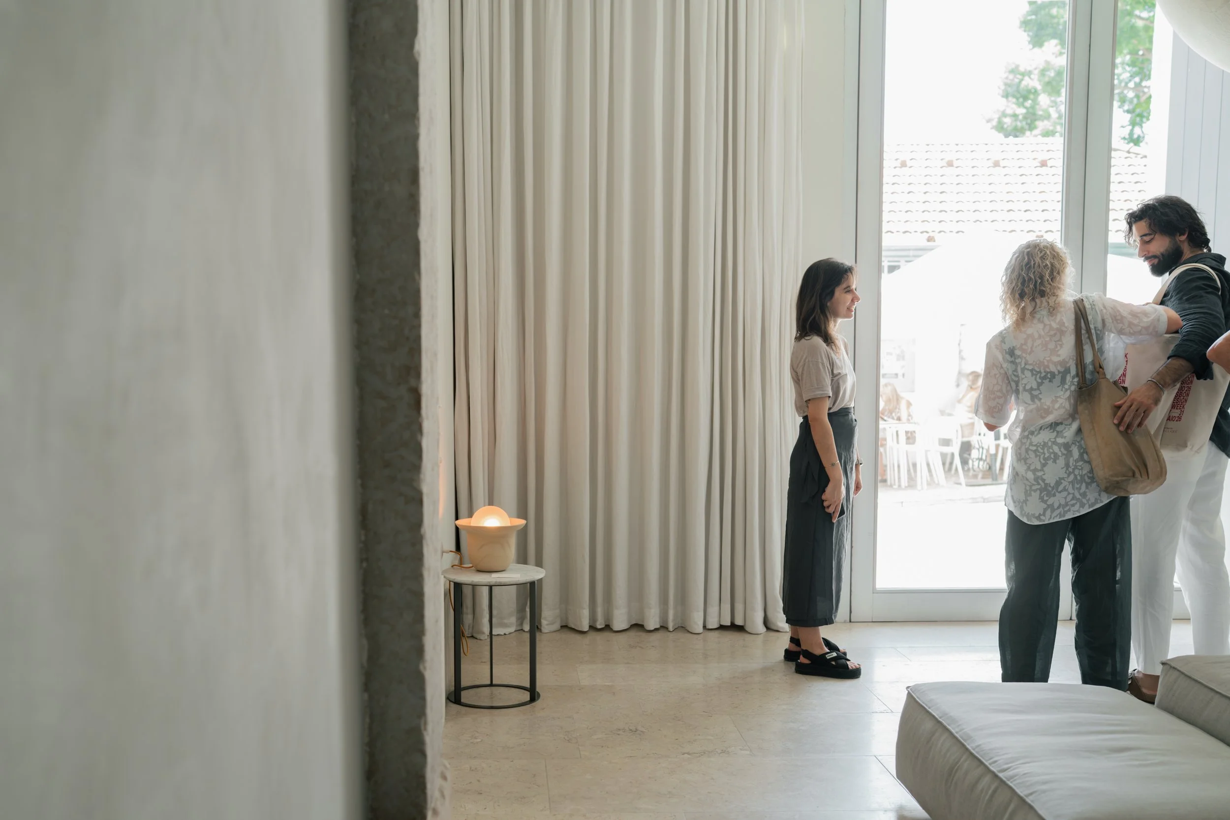 A group of four people standing near a large window in a well-lit room, engaging in a conversation. There is a small table with a lit lamp on it near the wall. The room has light-colored curtains and appears to be a modern, minimalist space with outd