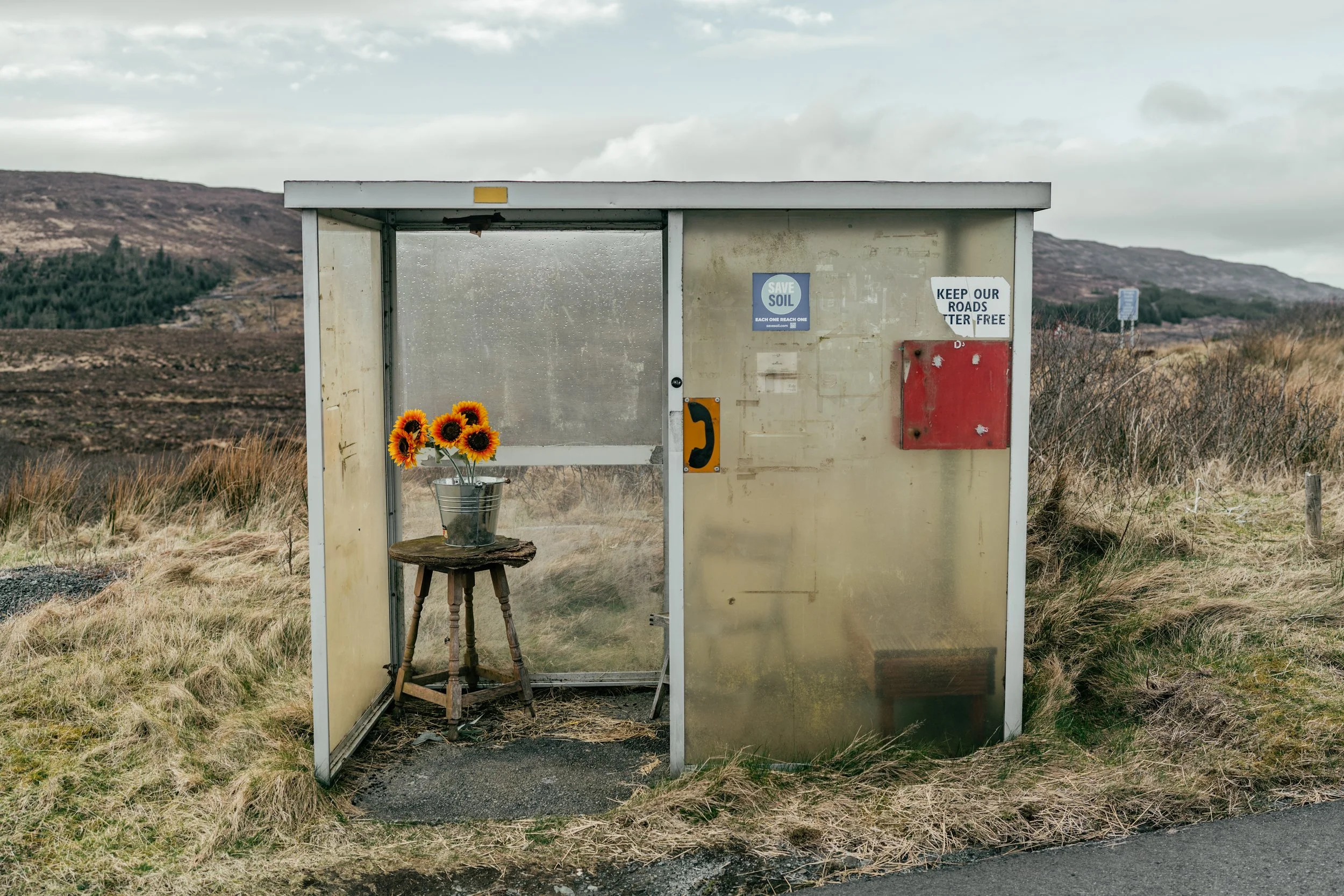 A small bus shelter in a rural area with a sunflower arrangement in a silver bucket on a wooden stool inside. The shelter has signs about soil conservation and road safety, with a landscape of hills and grasses in the background.