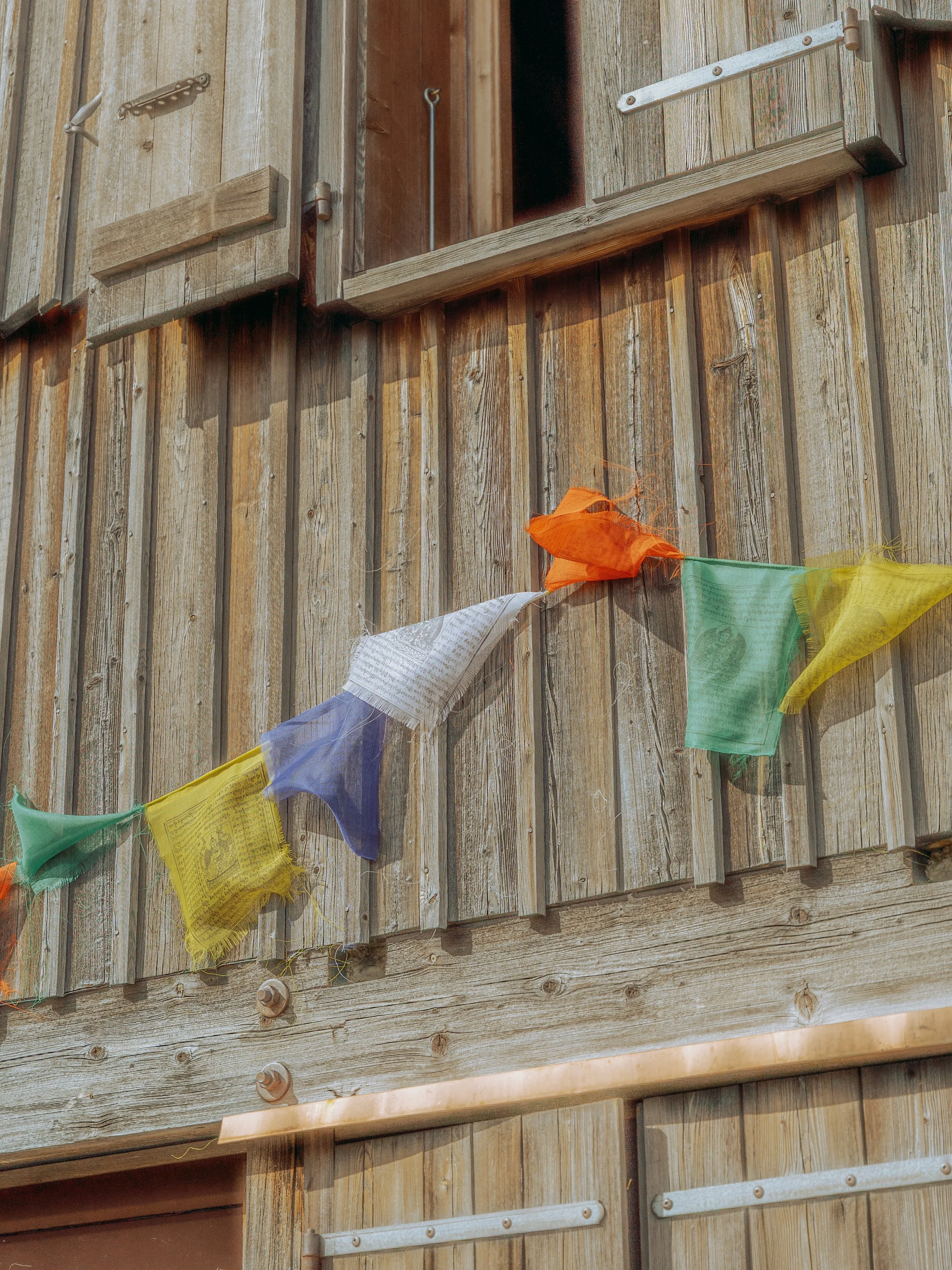 Close-up of a weathered wooden door with a display of colorful prayer flags hanging across it. The door features metal hinges and bolts, and the prayer flags are made of lightweight fabric, frayed at the edges.