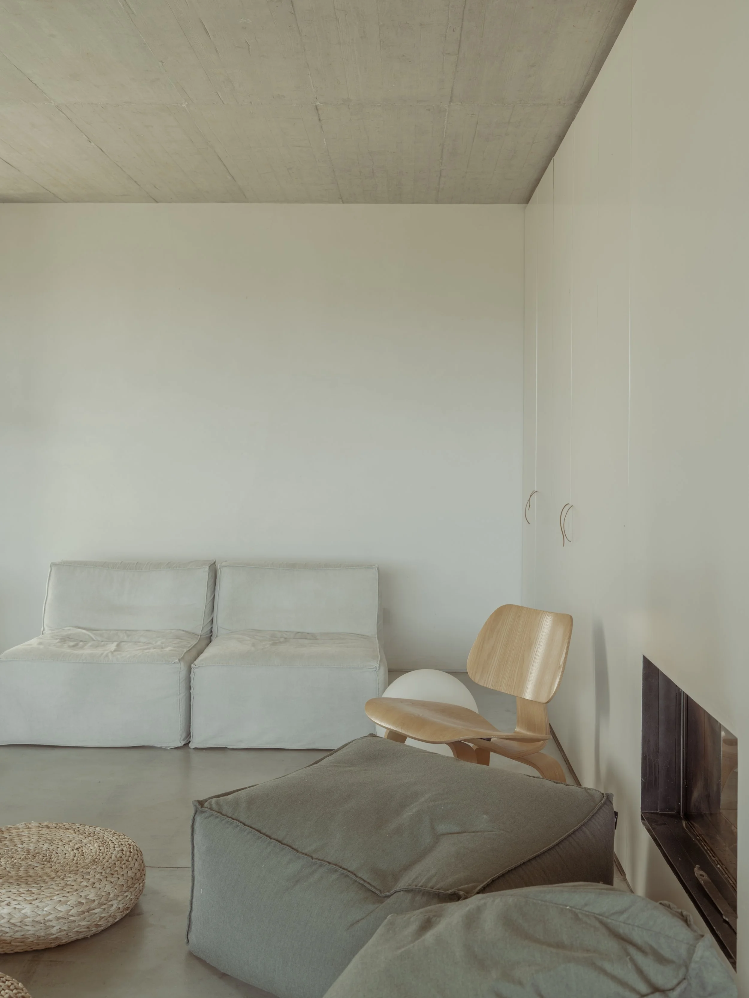 Living room with white sofa, gray armchair, wooden chair, TV, and woven ottomans, with a concrete ceiling and white walls.