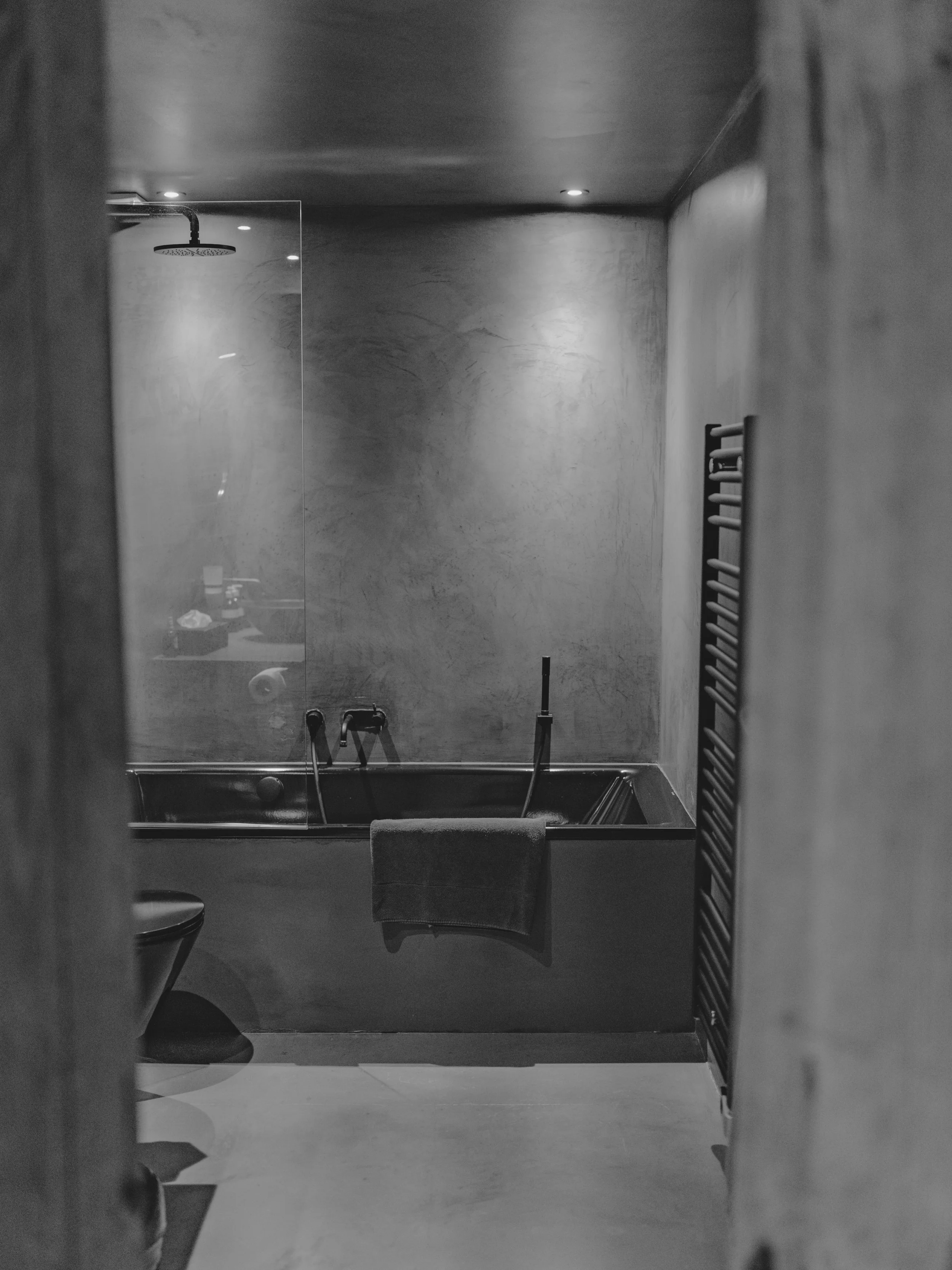 A black-and-white photo of a bathroom with a shower, a towel hanging on a ladder, a toilet, and other bathroom fixtures seen through a mirror reflection.