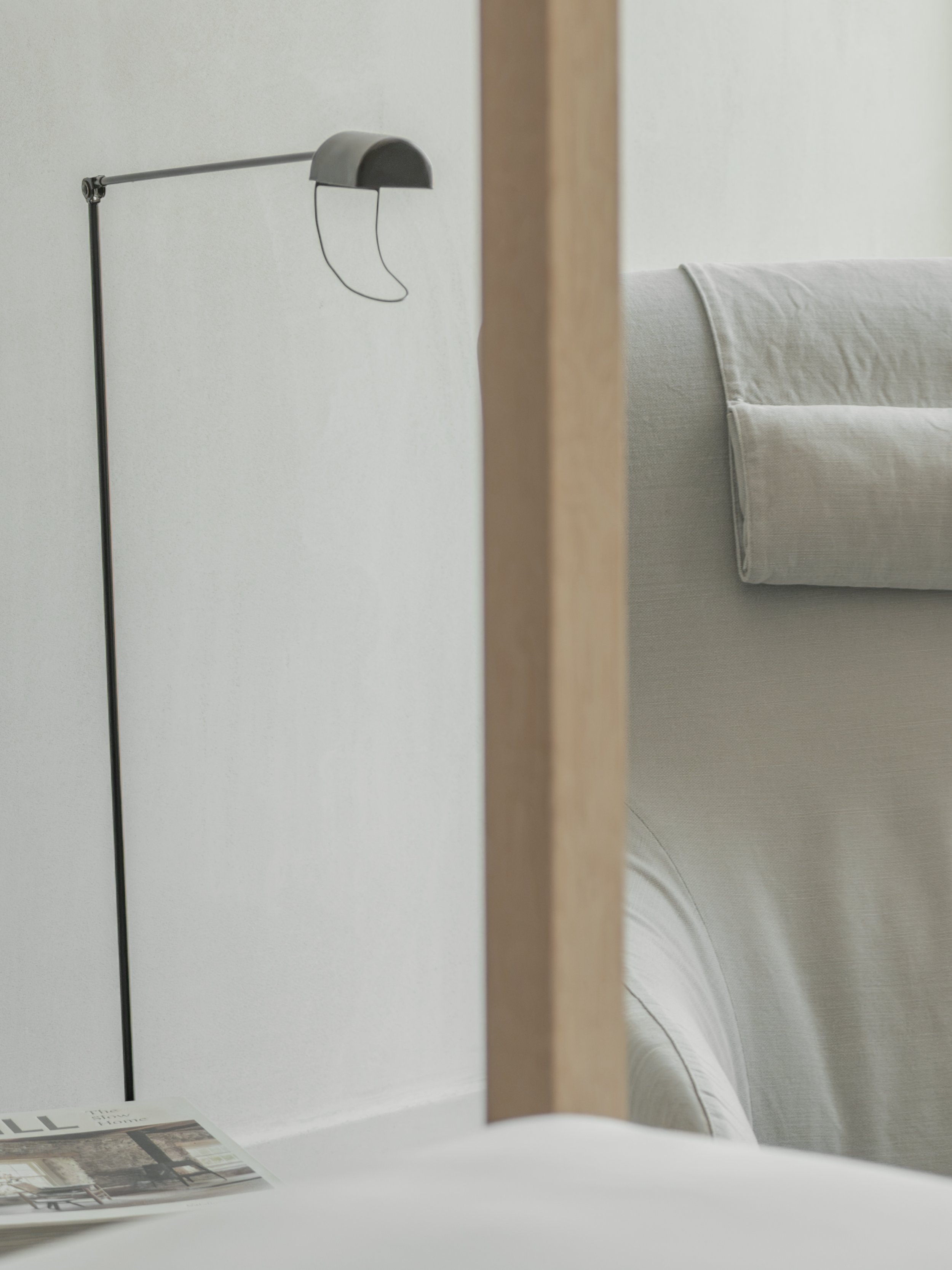 Close-up of a modern, minimalist desk lamp with a curved, metallic shade and thin wire arm, viewed through a paper or magazine on a white surface in a light-colored room.