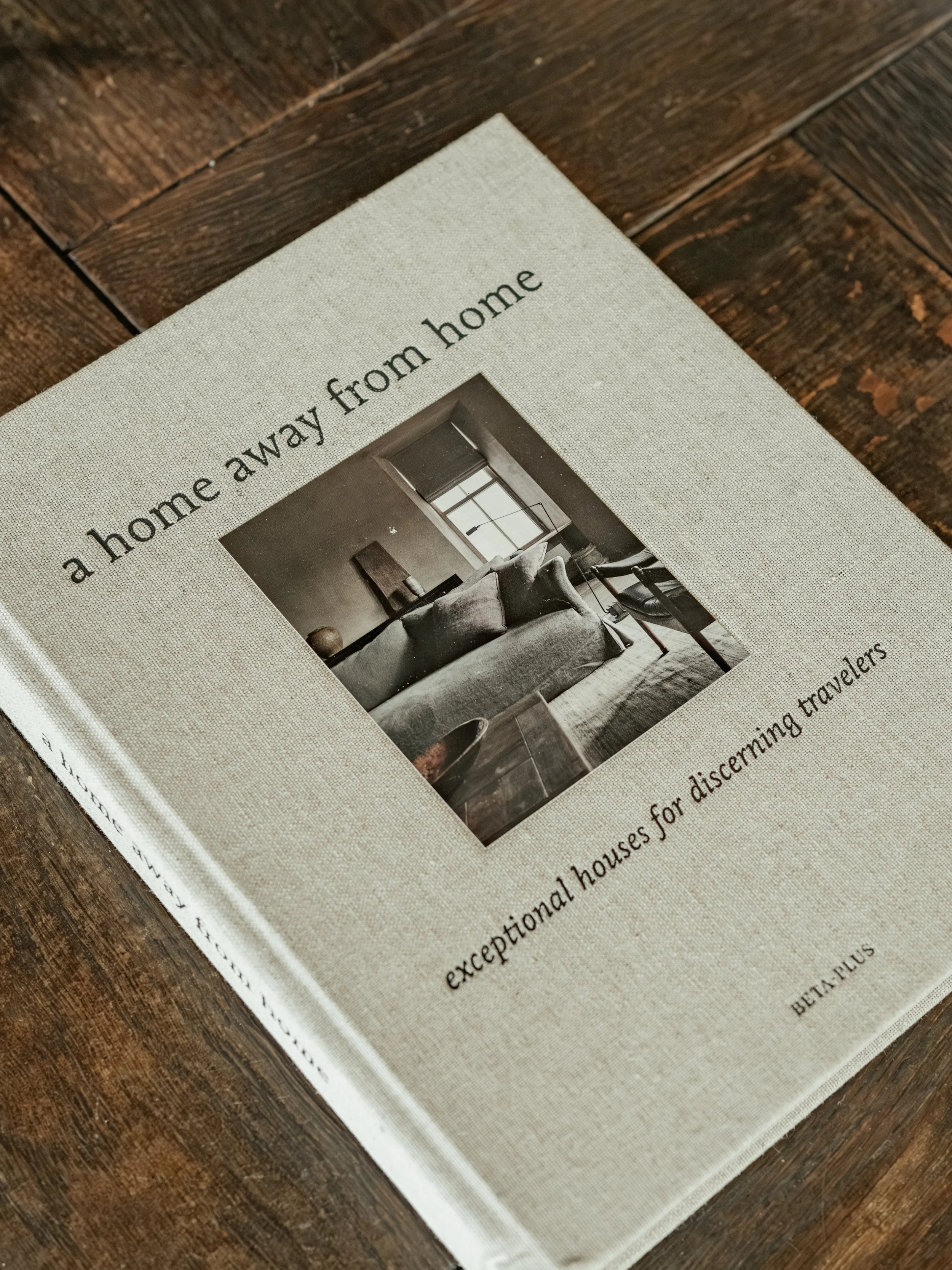 Book titled 'a home away from home' resting on a wooden surface, featuring a black and white photograph of a cozy living room with a couch and window on the cover.