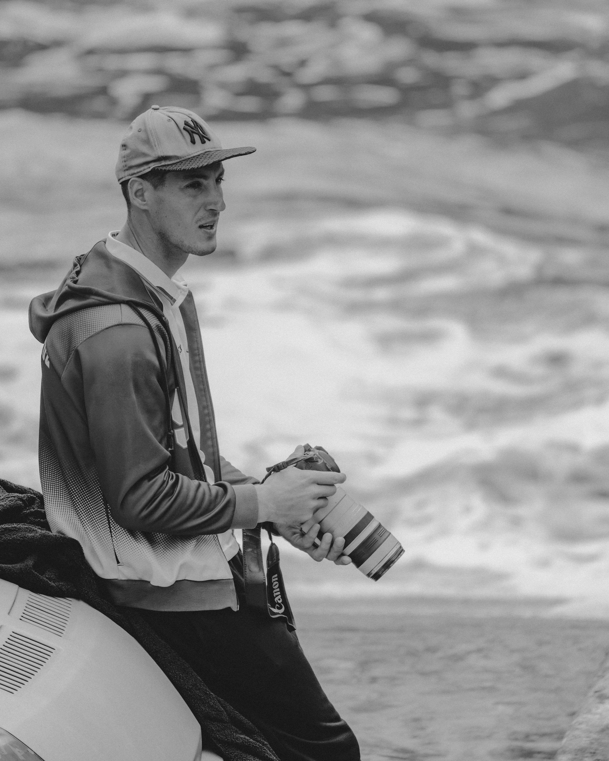 A man stands by the ocean holding a camera with a telephoto lens, wearing a New York Yankees cap and a jacket, with waves in the background.