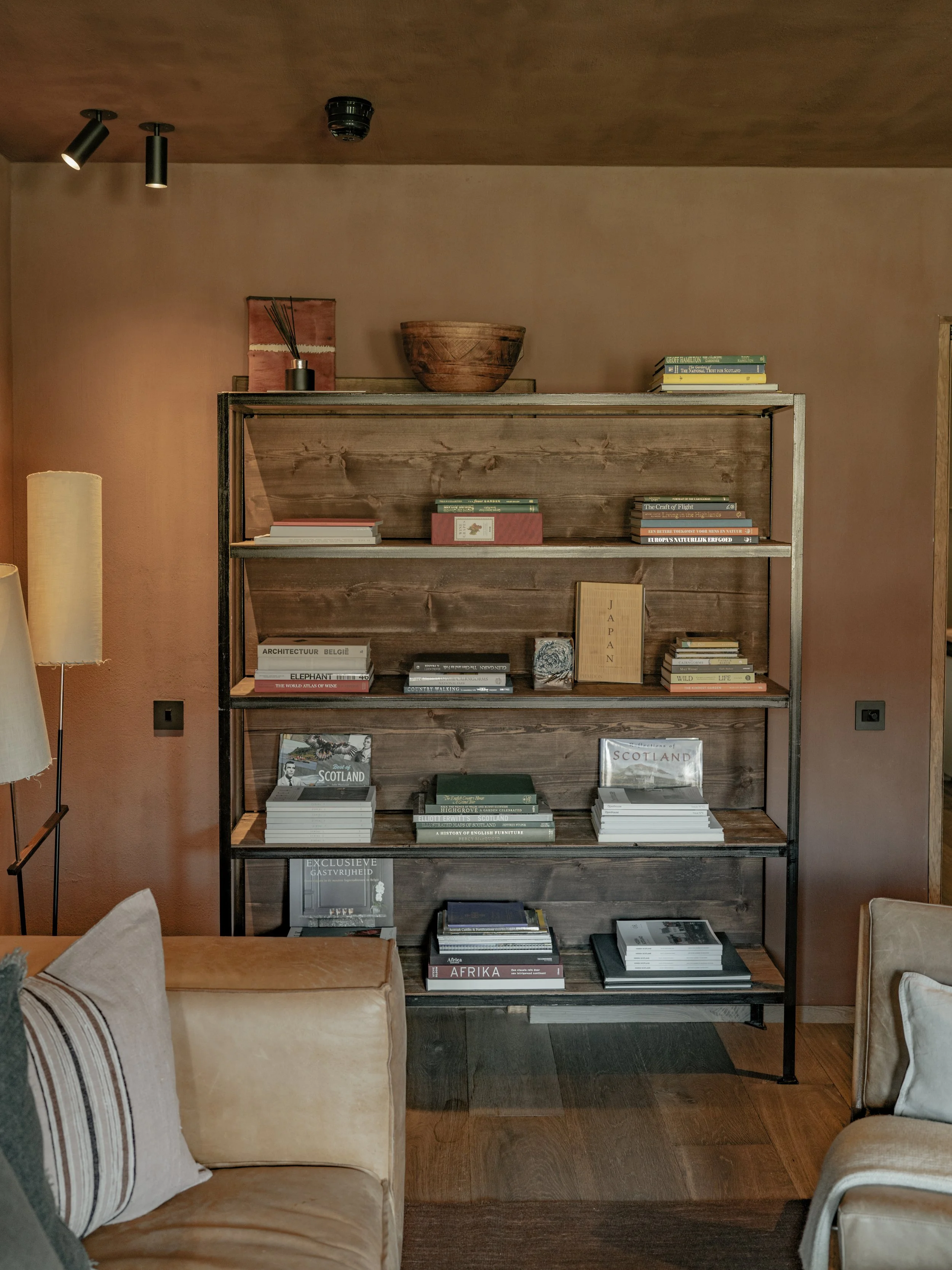 A wooden bookcase filled with various books on topics such as architecture, Scotland, and Africa, set against a warm-colored wall with a brown ceiling. Decorative items like a wooden bowl and small artworks are on top of the bookcase.
