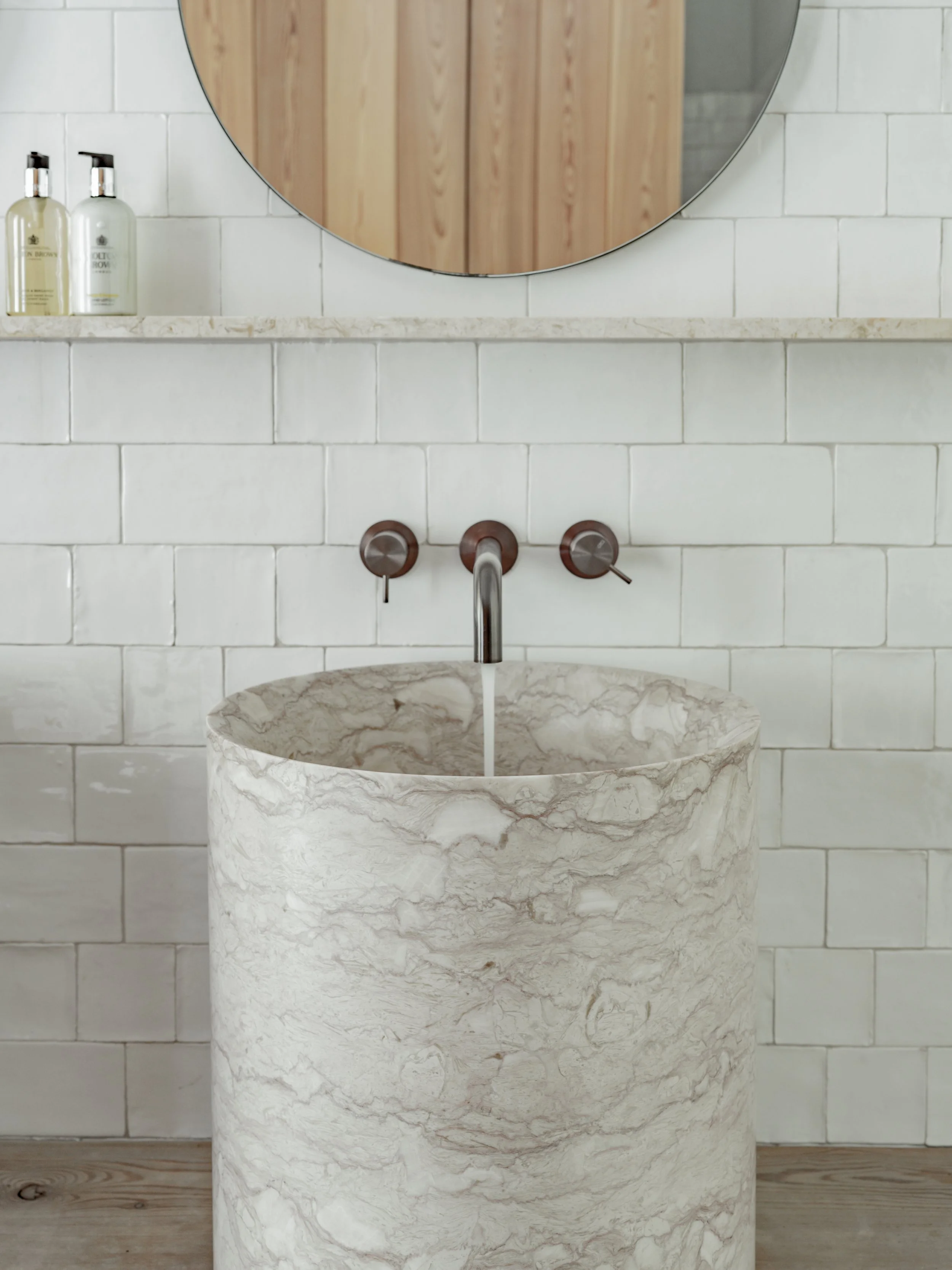 A restroom sink made of light-colored marbled stone with a round basin, mounted against a white tiled wall. Above the sink, there is a reflective round mirror. To the left, on a shelf, are two bottles of liquid soap or lotion. The faucet has a modern