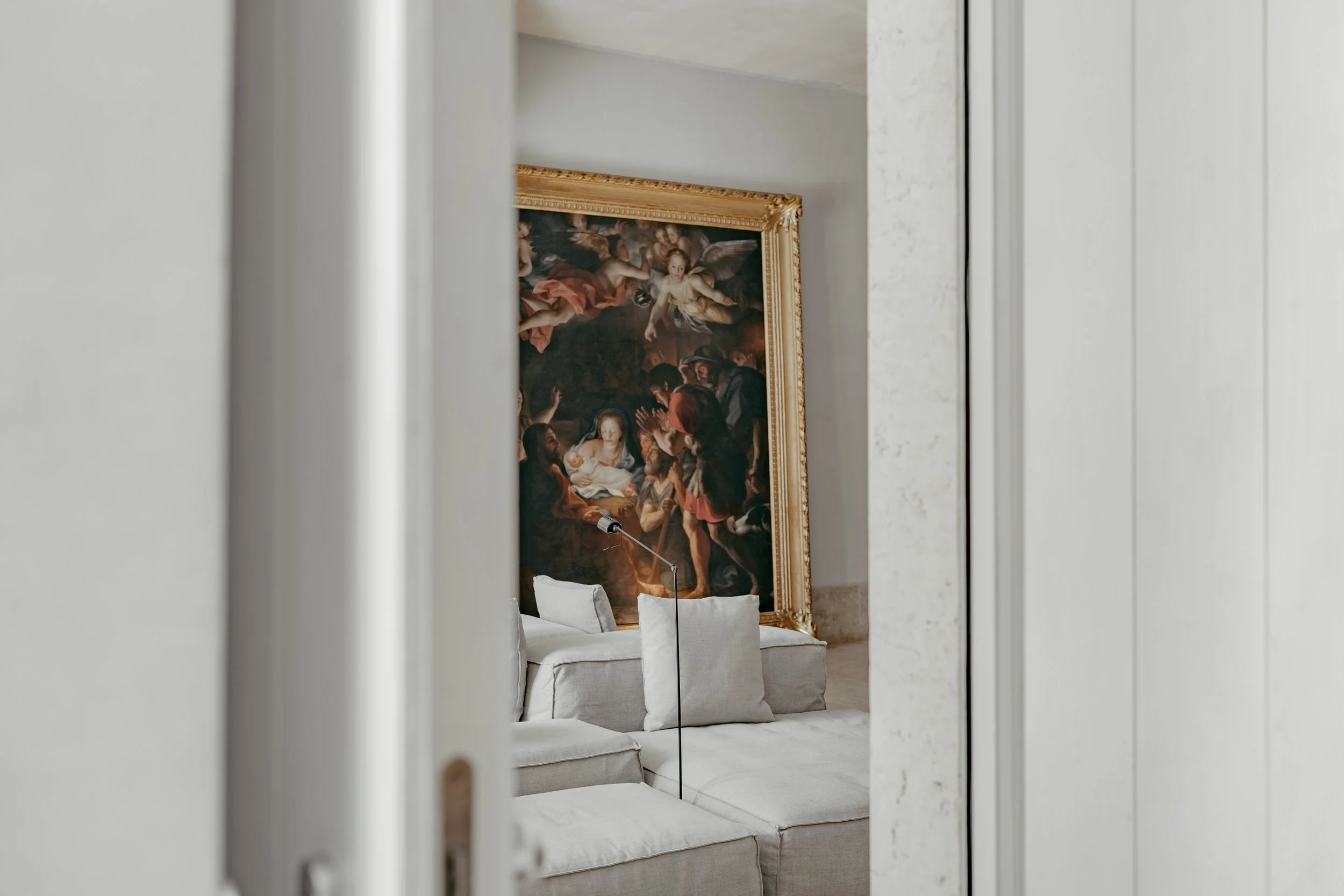 Partially visible classical painting with a gold frame, depicting a religious scene, on a white wall behind a white sofa in a well-lit room.