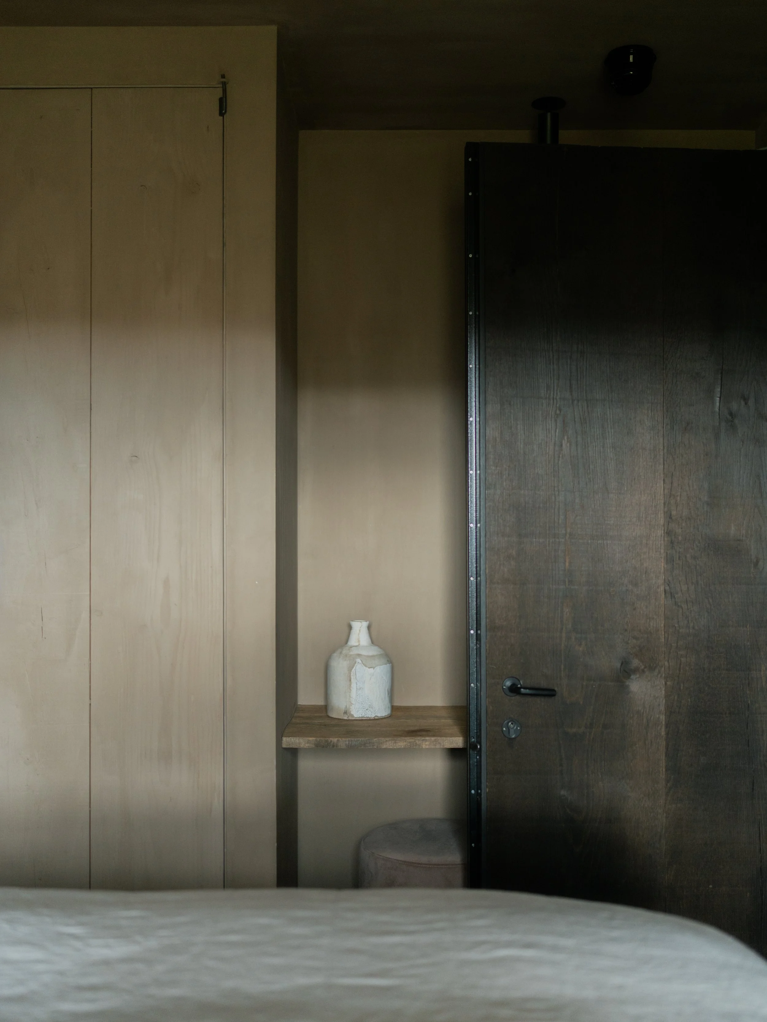 A partially opened wooden door reveals a beige wall with a small shelf holding a rustic white ceramic vase. To the left is a beige wardrobe with simple panel doors, and the floor is light-colored wood.