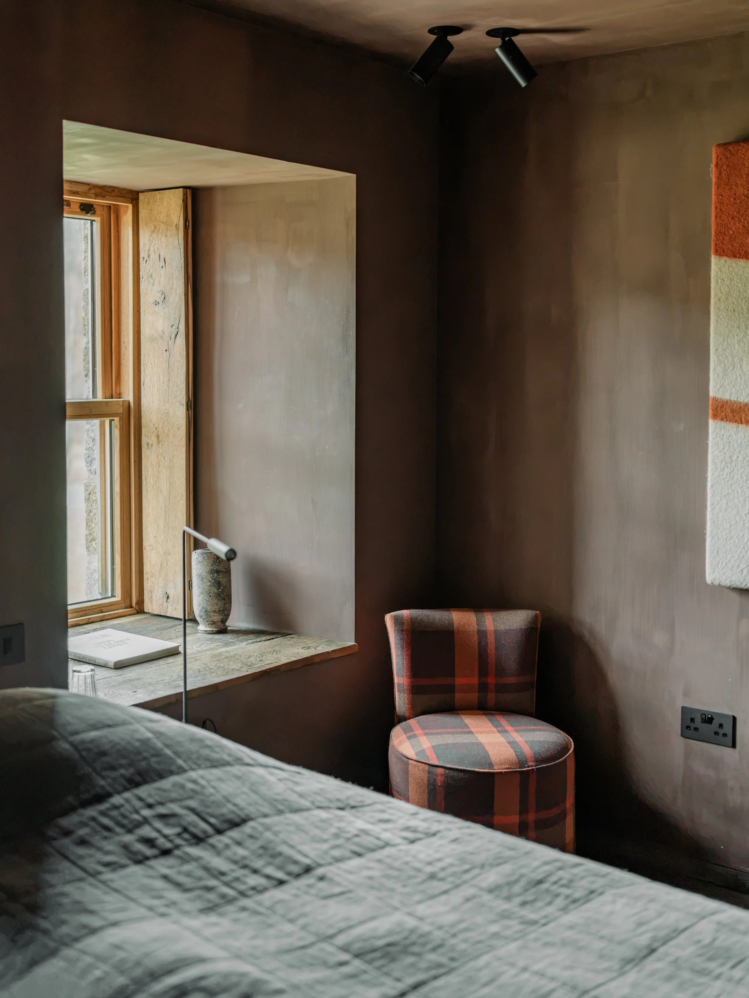A cozy bedroom corner with a bed in the foreground, a plaid upholstered chair, a window with natural light, a framed book, a tabletop lamp, and an electrical outlet along the wall.