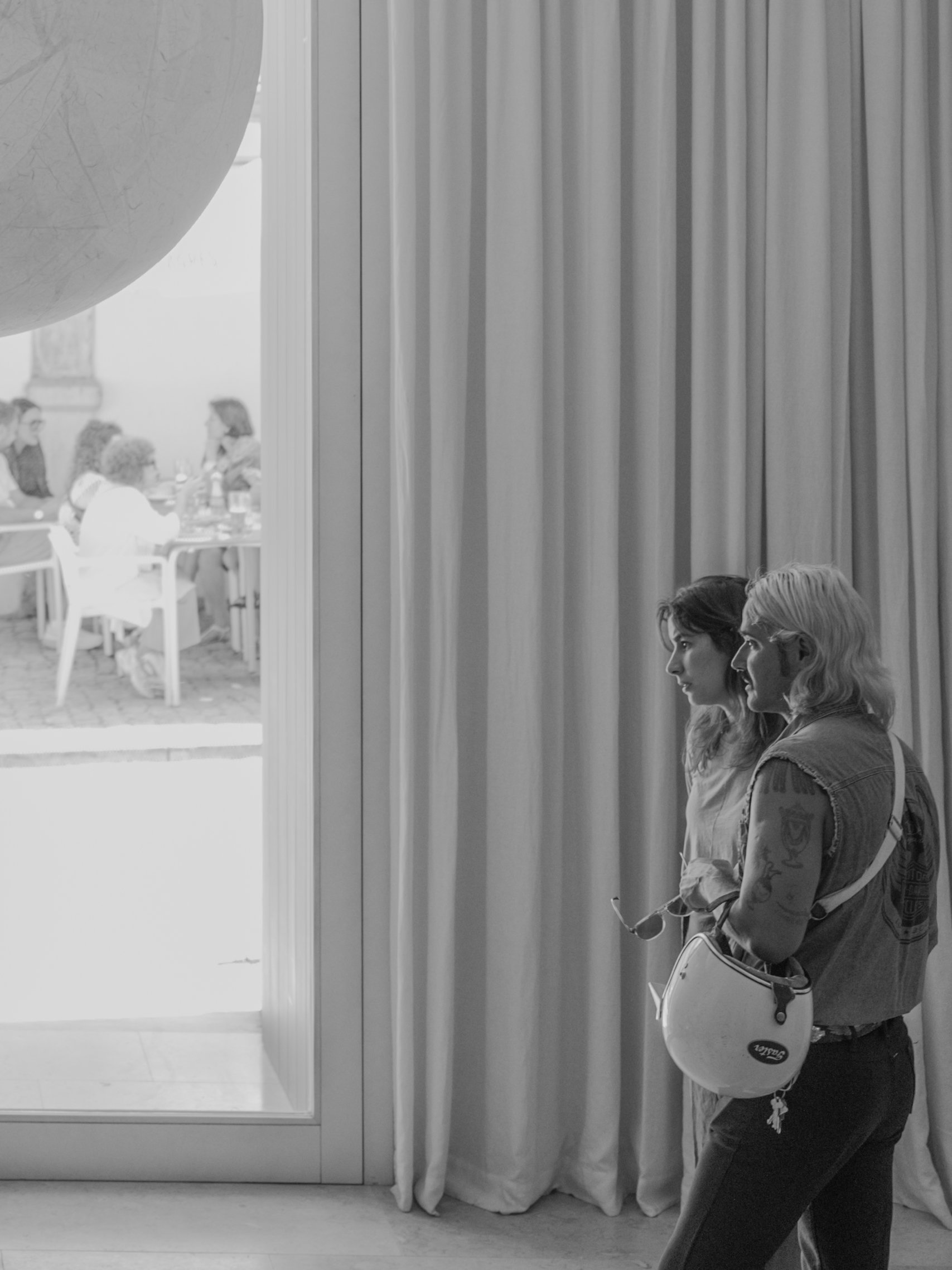 Young woman with tattoos holding a white helmet, standing near a window with curtains, with people sitting at tables visible in the background.