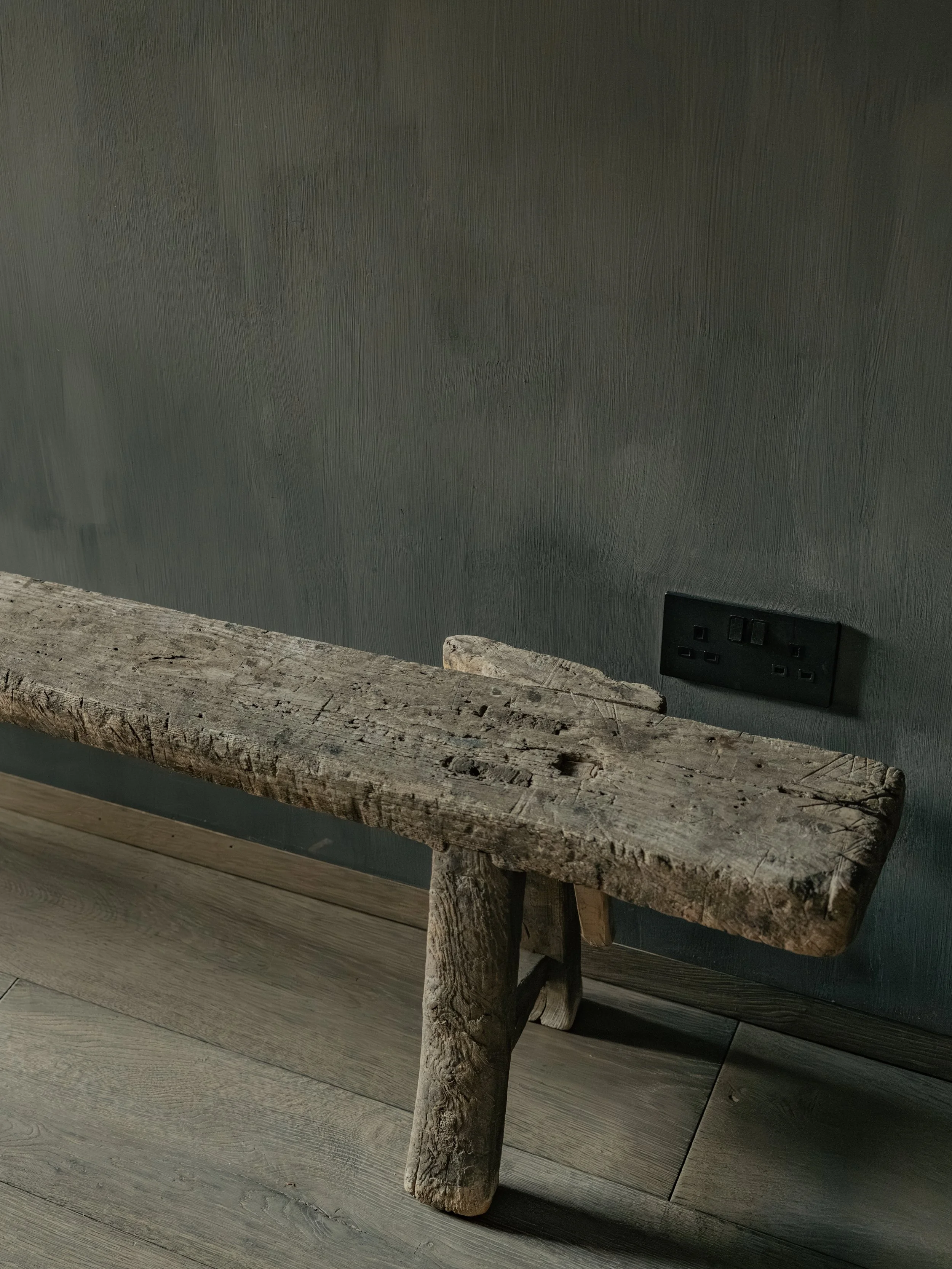 Close-up of a rustic wooden bench and a wall socket on a dark textured wall.