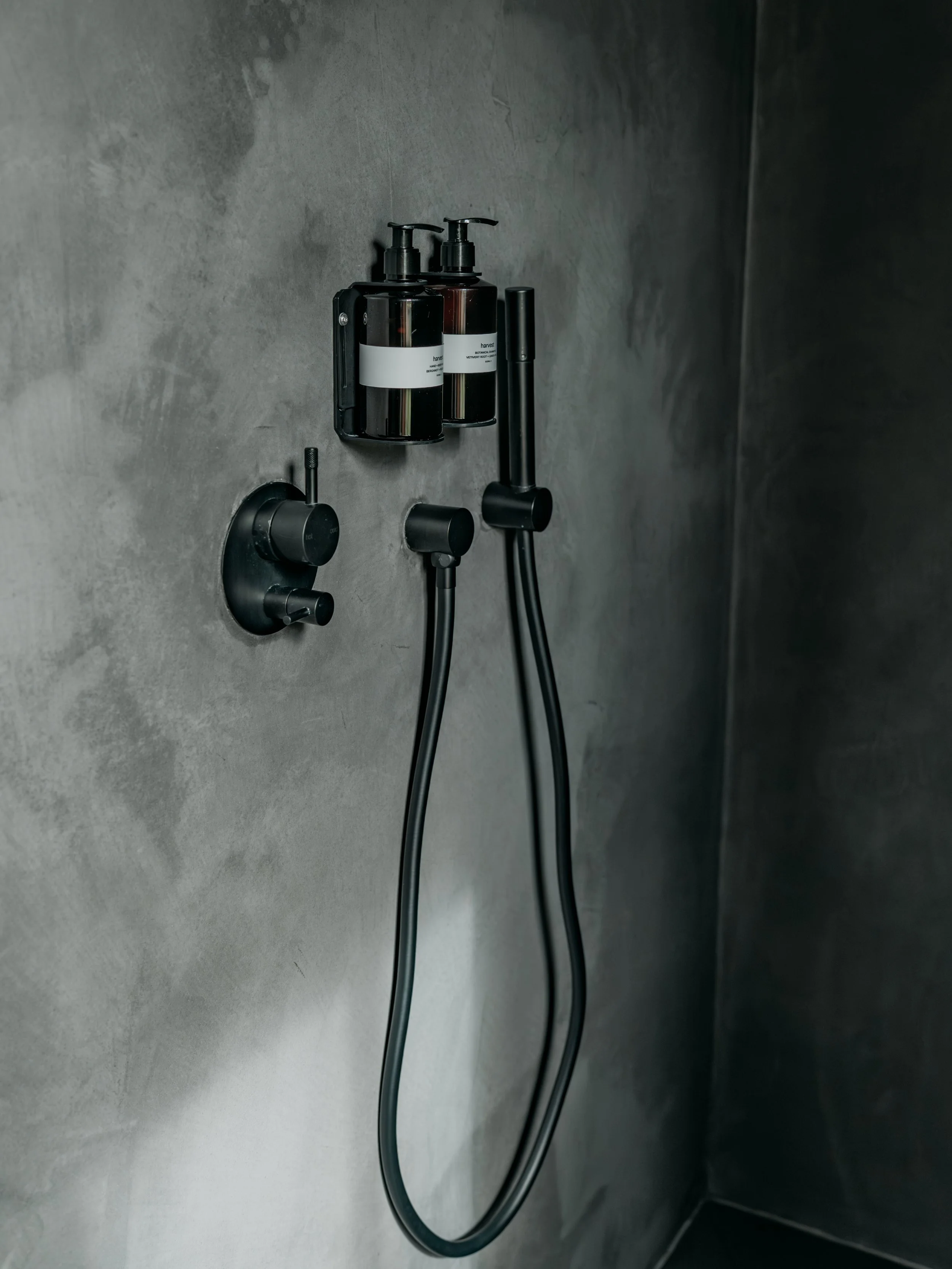 Close-up of two black soap or lotion bottles with pump dispensers, mounted on a wall in a bathroom or shower stall, with one black handheld showerhead and a control knob below.